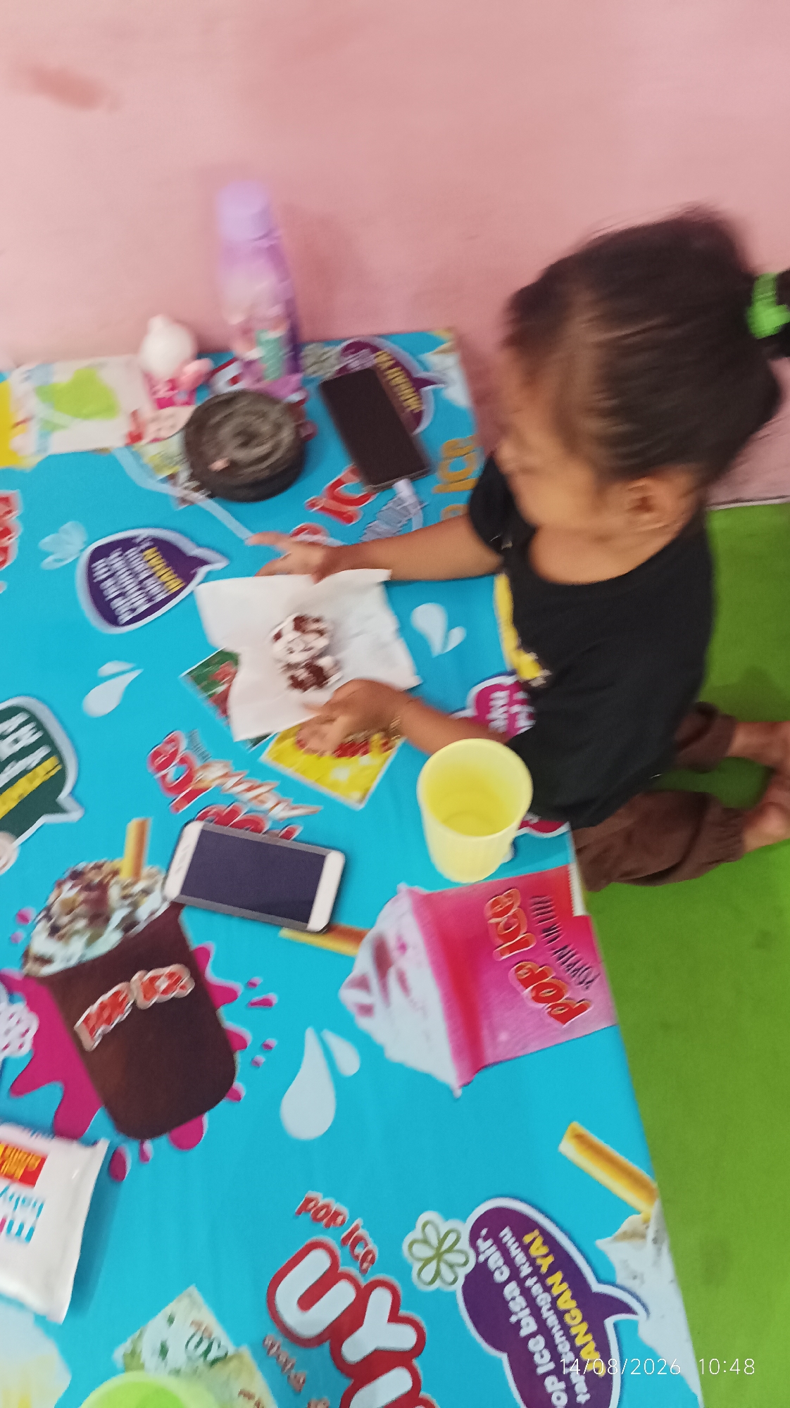 A child is sitting at a table covered with a blue tablecloth featuring a pattern of ice cream and text. The child is wearing a black t-shirt and brown pants, and is holding a white piece of paper with what appears to be a small ice cream on it. To the child's right is a yellow cup. On the table, there are also two mobile phones, one of which is in front of the child. In the background, there is a pink wall and a green surface on the floor. The image appears to have been taken indoors, possibly in a residential setting in Tasikmalaya, Indonesia, given the timestamp "11/08/2026 10:48" which indicates the date and time the photo was taken.