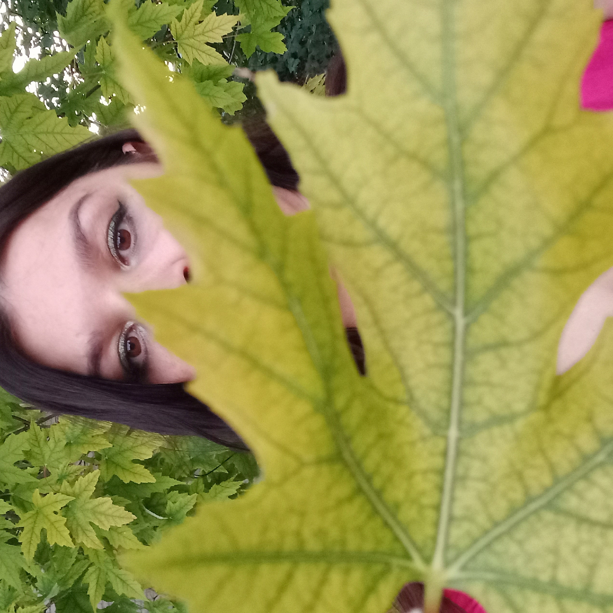 A woman with dark hair and green eyeshadow is partially obscured by a large, yellow-green leaf in the foreground. Her eyes, nose, and part of her forehead are visible through a gap in the leaf. She is positioned centrally in the frame, with a blurred background of green and yellow foliage, suggesting an outdoor setting. The lighting is natural, with some bright spots indicating sunlight filtering through the leaves. The scene is likely captured in Berezhynka, Ukraine, given the foliage which is characteristic of the region.