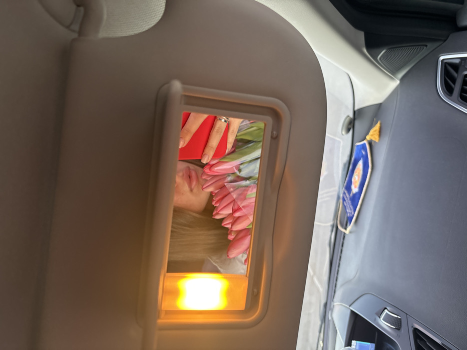 The image depicts the interior of a vehicle, specifically from the passenger's perspective looking towards the driver's side. The overhead sun visor is lowered, revealing an illuminated vanity mirror. Reflected in this mirror is the lower portion of a female's face, primarily her lips and chin, partially obscured by a bouquet of pink tulips wrapped in clear plastic. Her right hand, adorned with a silver ring on the ring finger, is visible holding a red smartphone, suggesting she is taking a selfie using the mirror. An amber-colored light fixture, part of the vanity mirror assembly, is illuminated on the left side of the mirror.
Below the sun visor, the dark gray dashboard of the car is visible, featuring air vents and what appears to be a digital display screen on the left. On the dashboard, a blue fabric face mask with a white design (possibly a sun or eye symbol) and white trim is laid flat. To its right, a small yellow or gold decorative tassel is present. The specific location within the city of Slavyansk-na-Kubani cannot be determined from the provided image beyond the internal setting of a car.