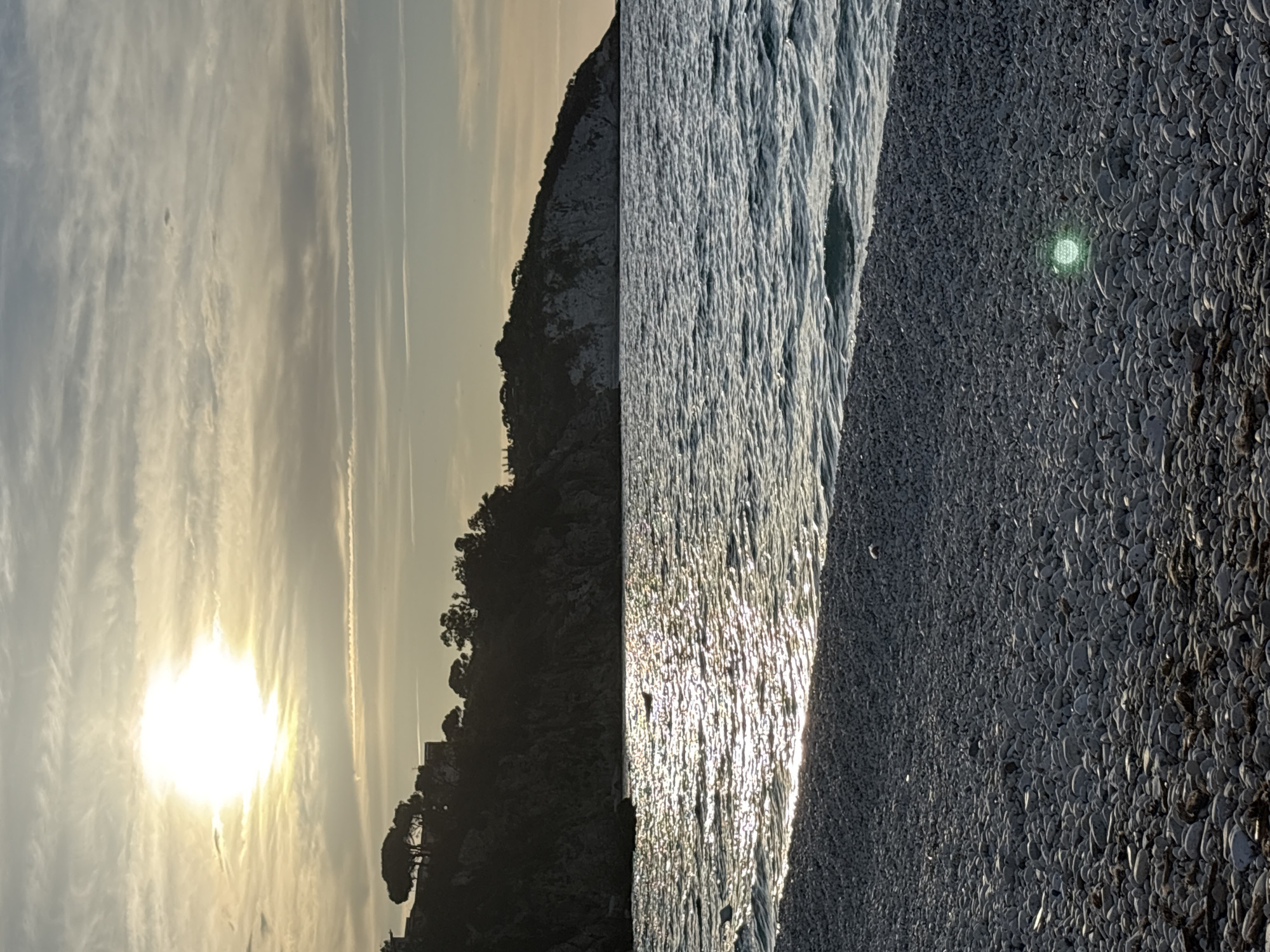 The image displays a serene coastal landscape captured during either sunrise or sunset, set in Portoferraio, Italy. The bright, low-lying sun dominates the left side of the sky, its intense light diffused by soft, wispy clouds. Faint contrails are visible against the pale, warm-toned sky.
To the left, a dark silhouette of a rugged, tree-covered promontory or cliff extends into the frame, its features obscured by the backlighting from the sun. The sea occupies the middle ground, showcasing a dazzling display of sunlight reflecting off its rippled surface, creating shimmering vertical streaks of light.
On the right, a dark pebble or shingle beach stretches into the foreground, with individual stones and shells clearly discernible, indicating a textured surface. A notable detail on the beach is a small, bright green, circular reflection or object visible in the mid-right. No people or visible text are present. The scene evokes a tranquil and atmospheric mood, characteristic of a Mediterranean coastal evening or morning.