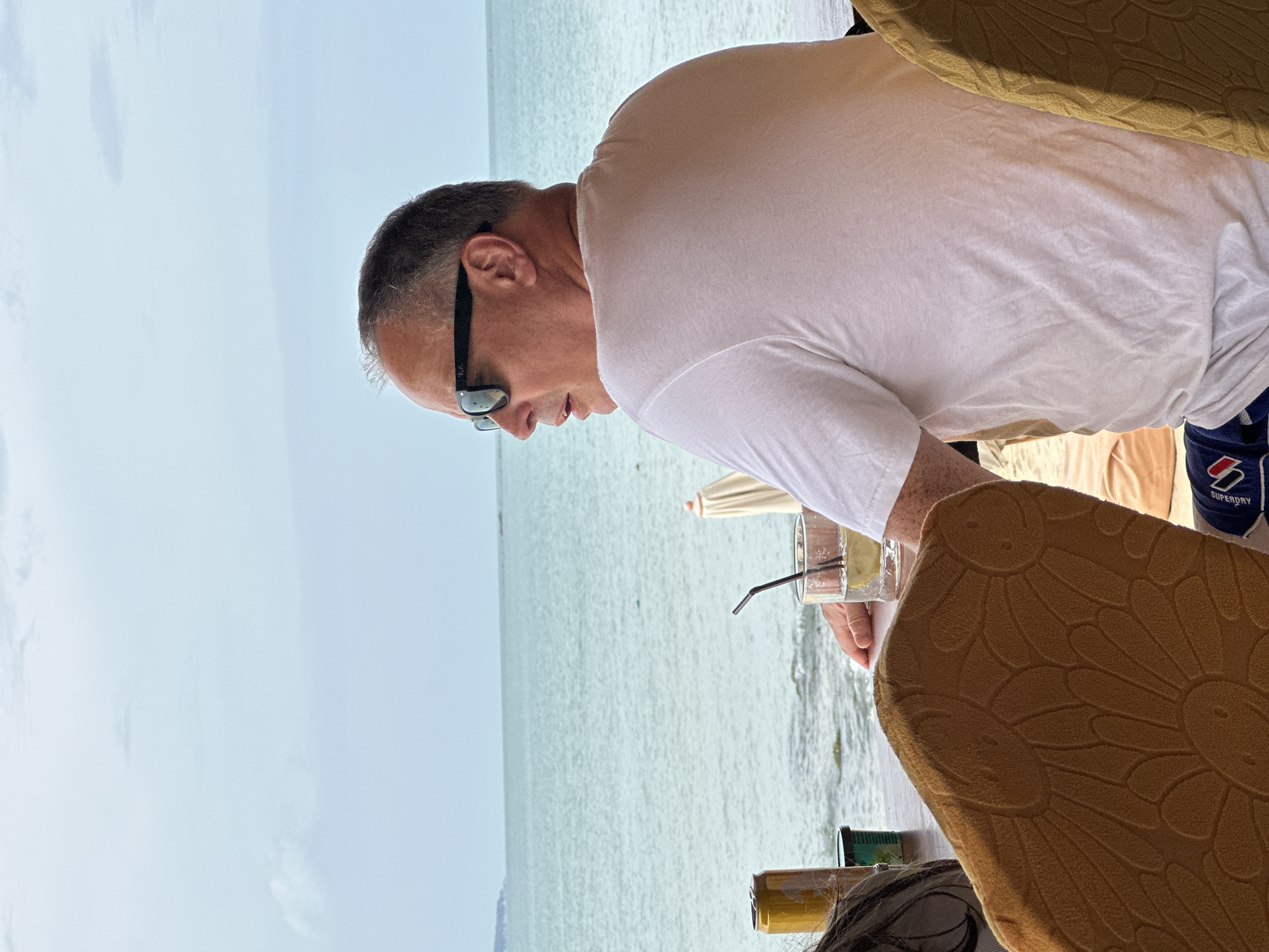 The image depicts a middle-aged Caucasian man with short, graying hair and sunglasses sitting at a table by the sea. He is wearing a white t-shirt and blue and white patterned shorts. The man is looking to his right, away from the camera. On the table in front of him is a glass with a drink and a straw, and a can. In the background, the turquoise sea stretches to the horizon under a cloudy sky. The scene appears to be set in a beachside restaurant or bar in Ko Samui District, Thailand, suggested by the tropical setting. The man is seated in a chair with a patterned upholstery.