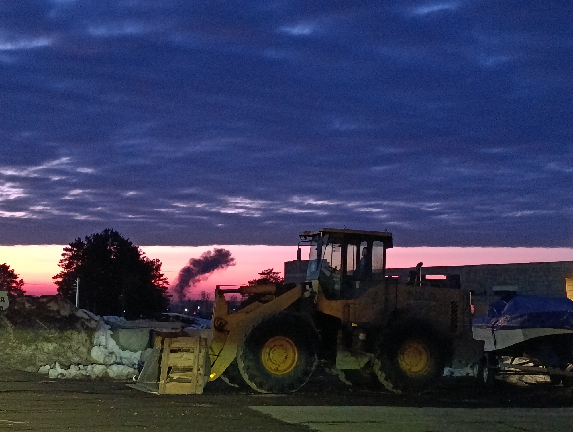 The image captures an outdoor scene at either dawn or dusk in Chaikovsky, Russia. The sky occupies the upper portion, displaying a gradient from deep blue and purple overhead to vibrant pink and orange at the horizon, with horizontal streaks of lighter clouds. The ground in the foreground is dark, uneven earth or gravel.
A large, yellow wheel loader is parked centrally in the mid-ground, oriented towards the left. Its front bucket contains a wooden pallet or crate. The machine appears weathered. To the left of the loader, a mound of dark earth or debris is partially covered with dirty snow or ice. Behind this mound, a cluster of dark evergreen trees is silhouetted against the bright horizon. A distinct plume of dark grey smoke rises vertically from behind these trees.
To the right of the loader, a light-colored building is visible. Adjacent to the building, a white boat on a trailer, covered with a blue tarp, is parked. No individuals are visible, and no active interactions or movements are occurring within the frame; all objects appear stationary.