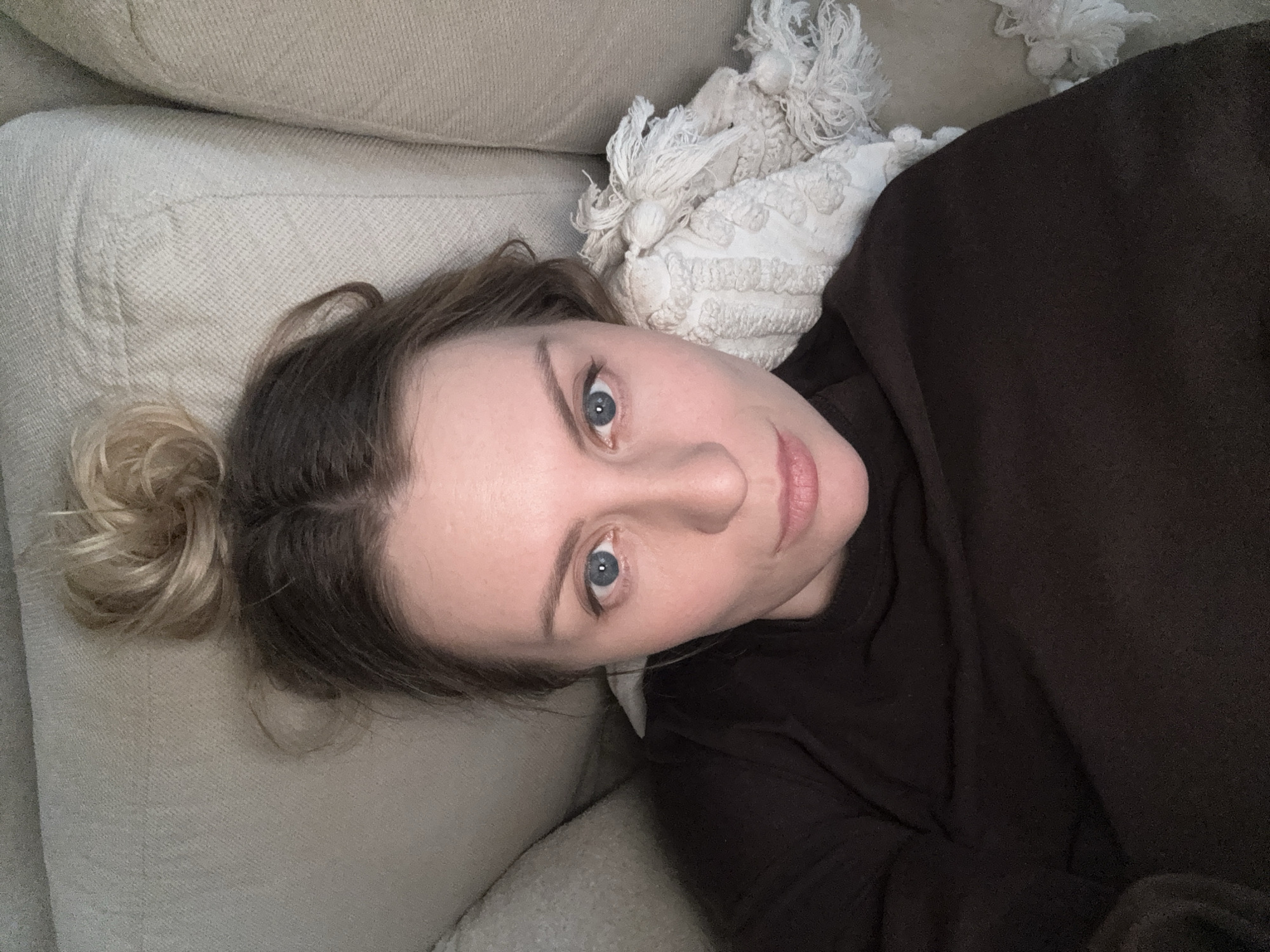 A single adult female is depicted in a close-up indoor portrait, likely a selfie. She has fair skin, blue eyes, and light brown hair with blonde highlights styled in a loose bun atop her head, revealing darker roots. Her expression is neutral, and she gazes directly into the camera. She wears a dark, possibly black or dark brown, long-sleeved garment. She is reclining against a light-colored, textured upholstered surface, consistent with a sofa or bed. A cream-colored, textured throw blanket or pillow with tassels is partially visible on the right side of the frame. The immediate environment consists solely of the upholstered furniture and textile. No visual information in the image allows for the determination of its specific location within the city of Moscow, Russia.
