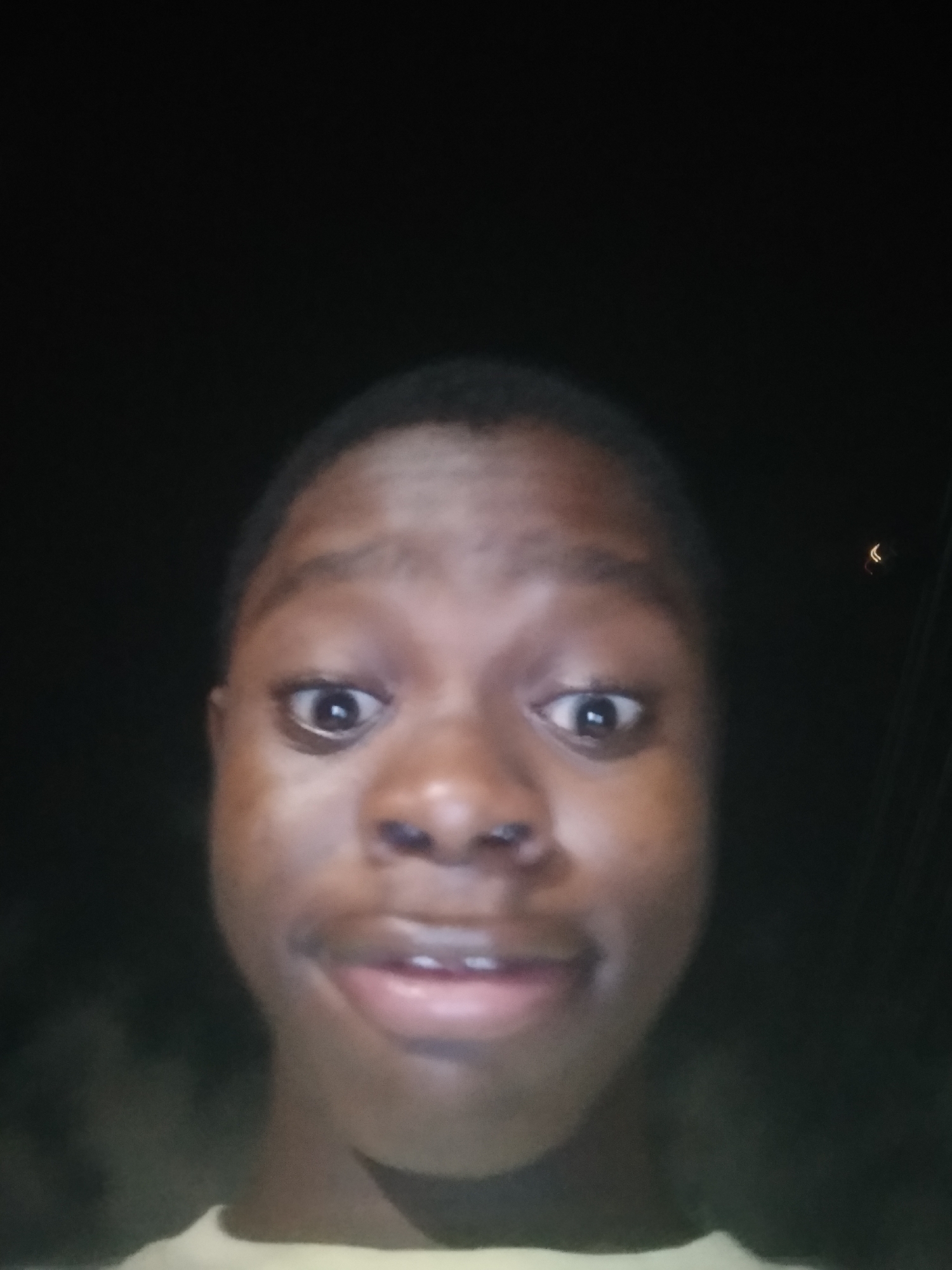 A person is taking a selfie at night. The person is dark-skinned with short hair, wide eyes, and a slightly open mouth. The background is dark, with some indistinct shapes possibly indicating foliage. A faint light source is visible in the upper right. The location is Unknown, Unknown.