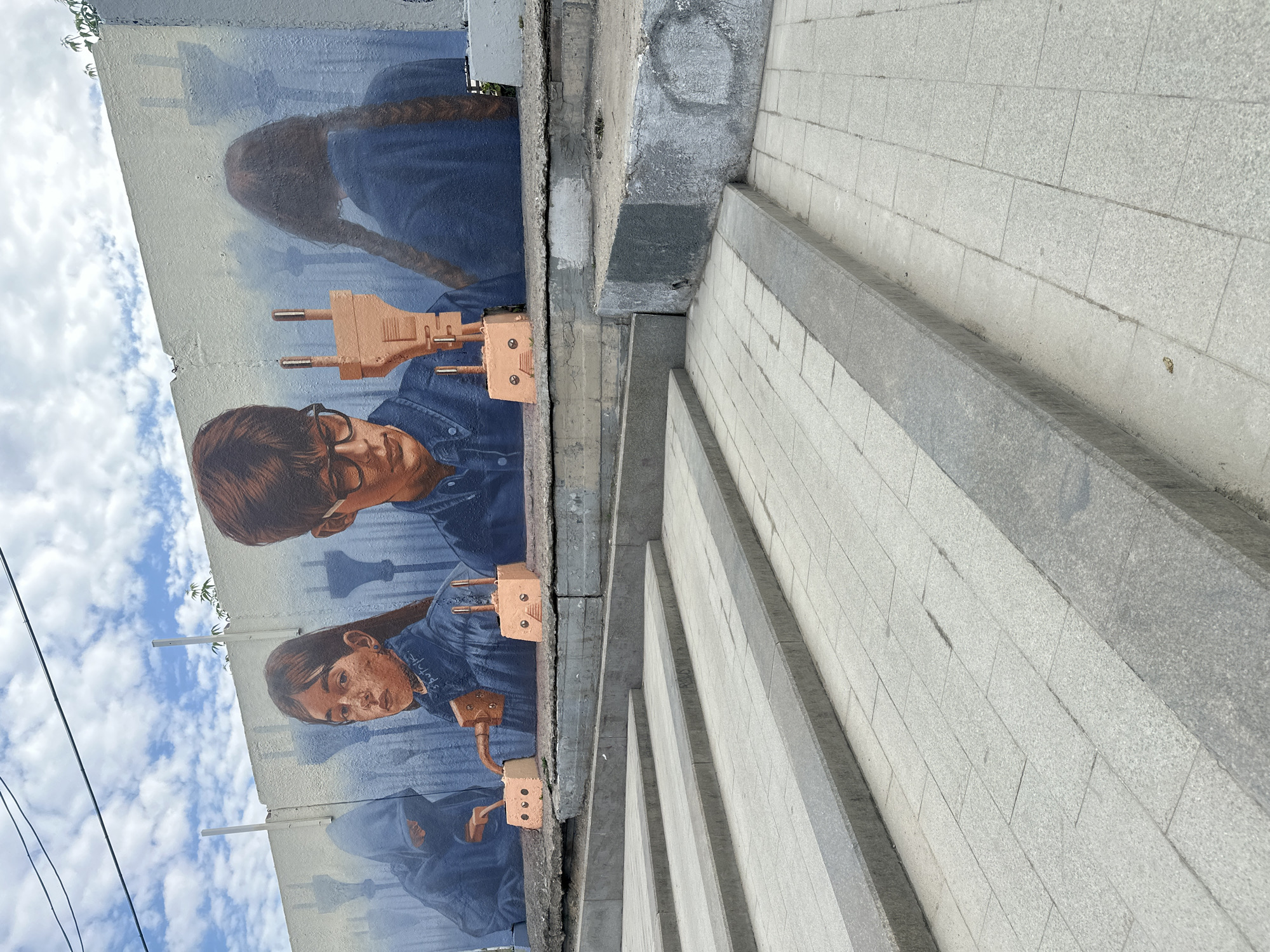 A street-level view captures a section of a pedestrian staircase adjacent to a wall adorned with a mural. The mural, seemingly influenced by surrealist or symbolic themes, features repeated images of children or young adults depicted with vintage-style electrical plugs extending from their heads or bodies. The subjects are painted with detailed faces and wearing denim clothing, set against a backdrop of a cloudy sky. The mural is affixed to a gray concrete wall, which also forms part of the staircase's retaining structure. The staircase is constructed of gray stone or concrete steps with visible mortar lines. The overall scene suggests an urban setting, possibly in Moscow, Russia, given the architectural style and the style of the mural.