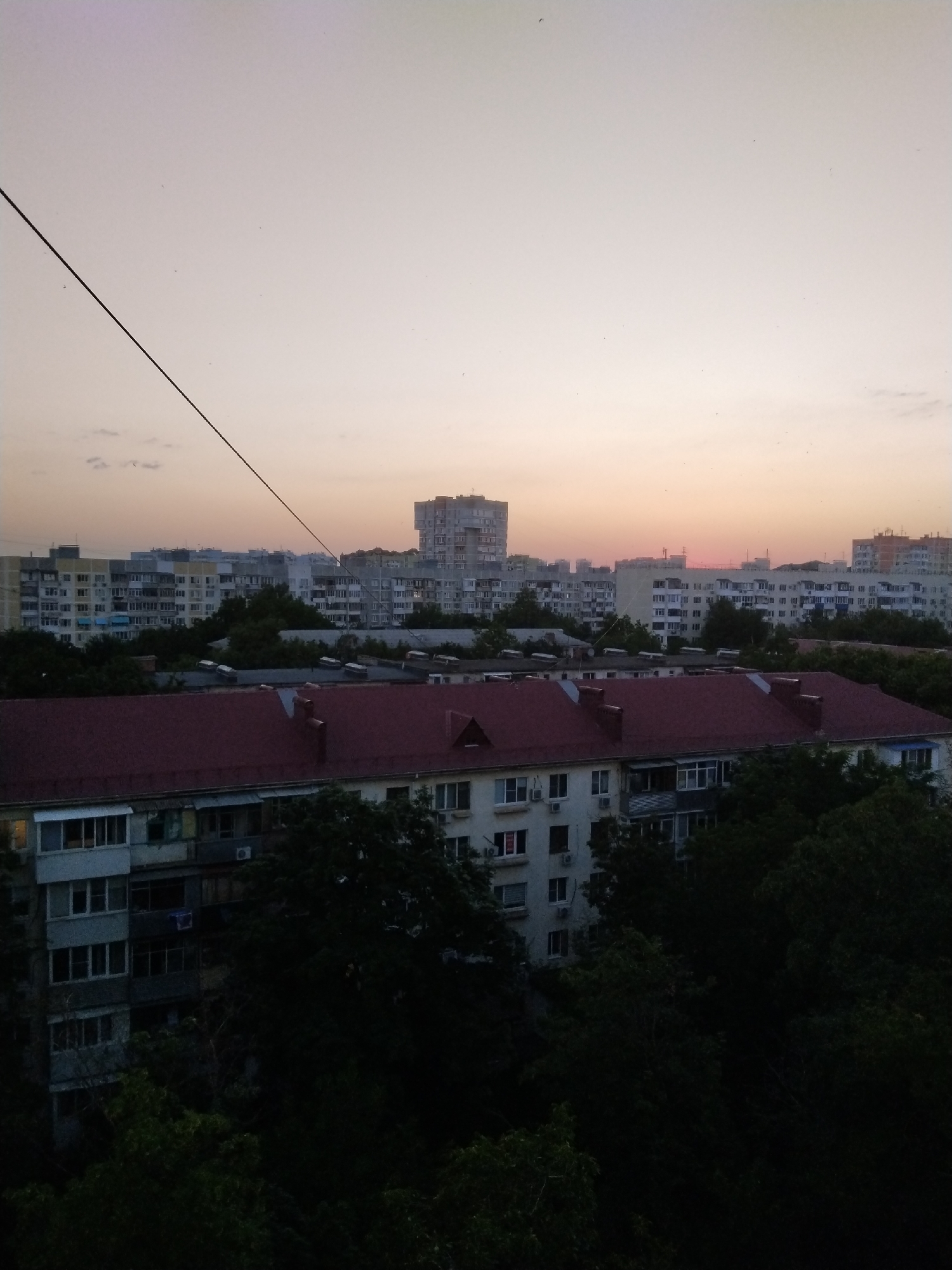 The image captures a panoramic view of a cityscape at dusk, likely from a high vantage point. The sky exhibits a gradient of soft peach and lavender hues, indicative of sunset. A solitary black wire, possibly an electrical or communication line, bisects the upper left quadrant of the image diagonally.
In the mid-ground, a densely packed residential area is visible. Numerous multi-story apartment buildings, characterized by their pale facades and uniform window patterns, dominate the scene. The architecture suggests a Soviet-era urban planning style, common in many Russian cities. Among these buildings, some appear to be taller and more modern, while others are lower and have distinctive tiled roofs, some in a deep red hue.
In the foreground, the rooftops of several lower buildings are prominent. One building in the immediate foreground has a dark red, pitched roof with visible architectural details such as dormer windows or vents. The lower sections of buildings are partially obscured by lush, dark green trees, indicating the presence of significant vegetation within the urban environment. The foliage suggests a temperate climate, and the dense trees might be a common feature of parks or residential courtyards.
The overall impression is one of a typical urban landscape in Russia during the evening, with a focus on residential architecture and the interplay of built structures with the natural elements of trees and the sky. The absence of visible people or activity suggests a quiet, observed moment. The scene is consistent with an urban environment found in cities like Krasnodar, Russia, which is known for its apartment blocks and green spaces.