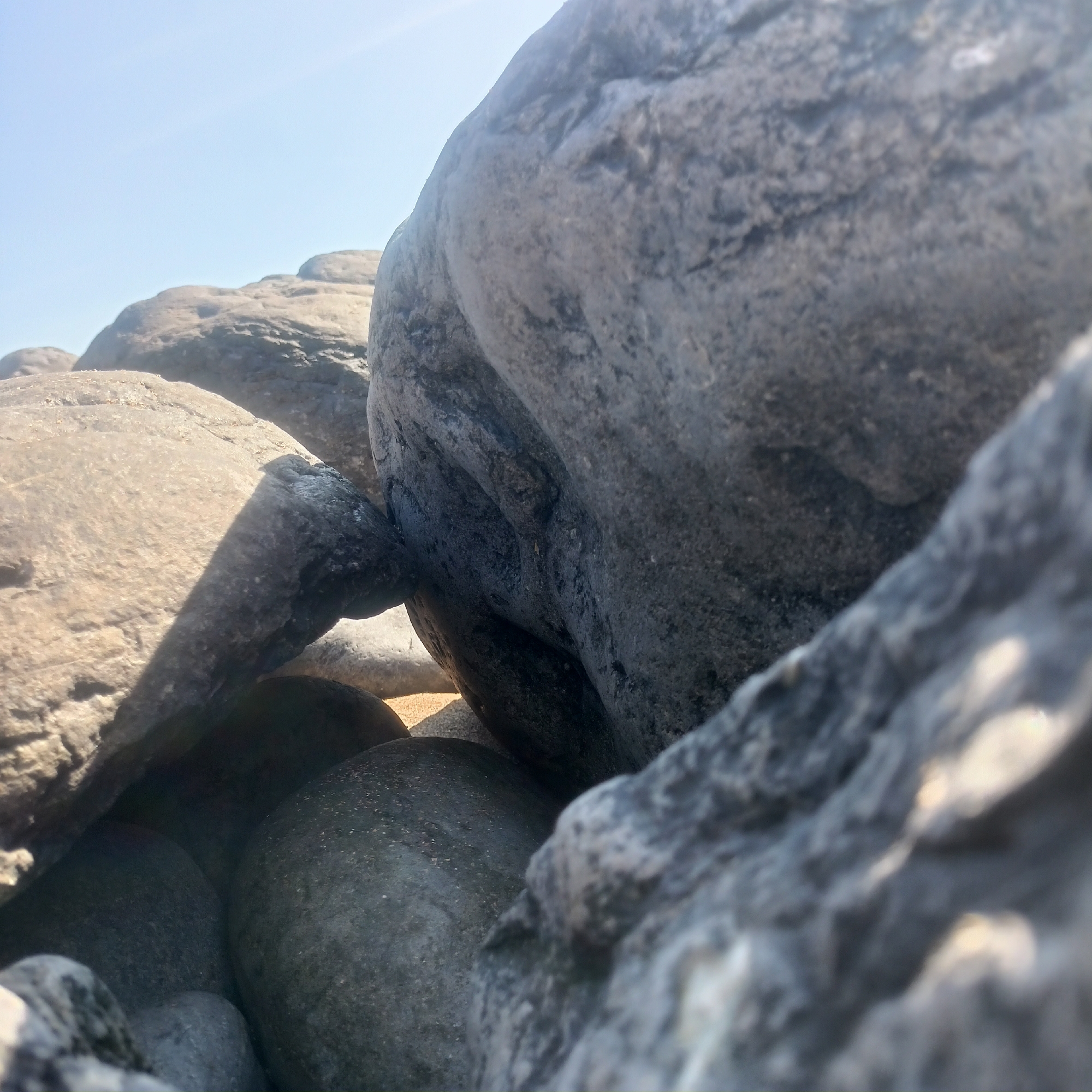 The image shows a close-up view of large, weathered rocks under a clear sky, possibly on a beach or coastline. The rocks vary in color from light brown to dark gray and exhibit textured surfaces. Some rocks are smooth and rounded, while others are more angular. The lighting suggests it is daytime, with sunlight casting shadows across the stones. The scene is devoid of any human presence or activity. The location is identified as Fanore, Ireland, suggesting this is a natural outdoor landscape.