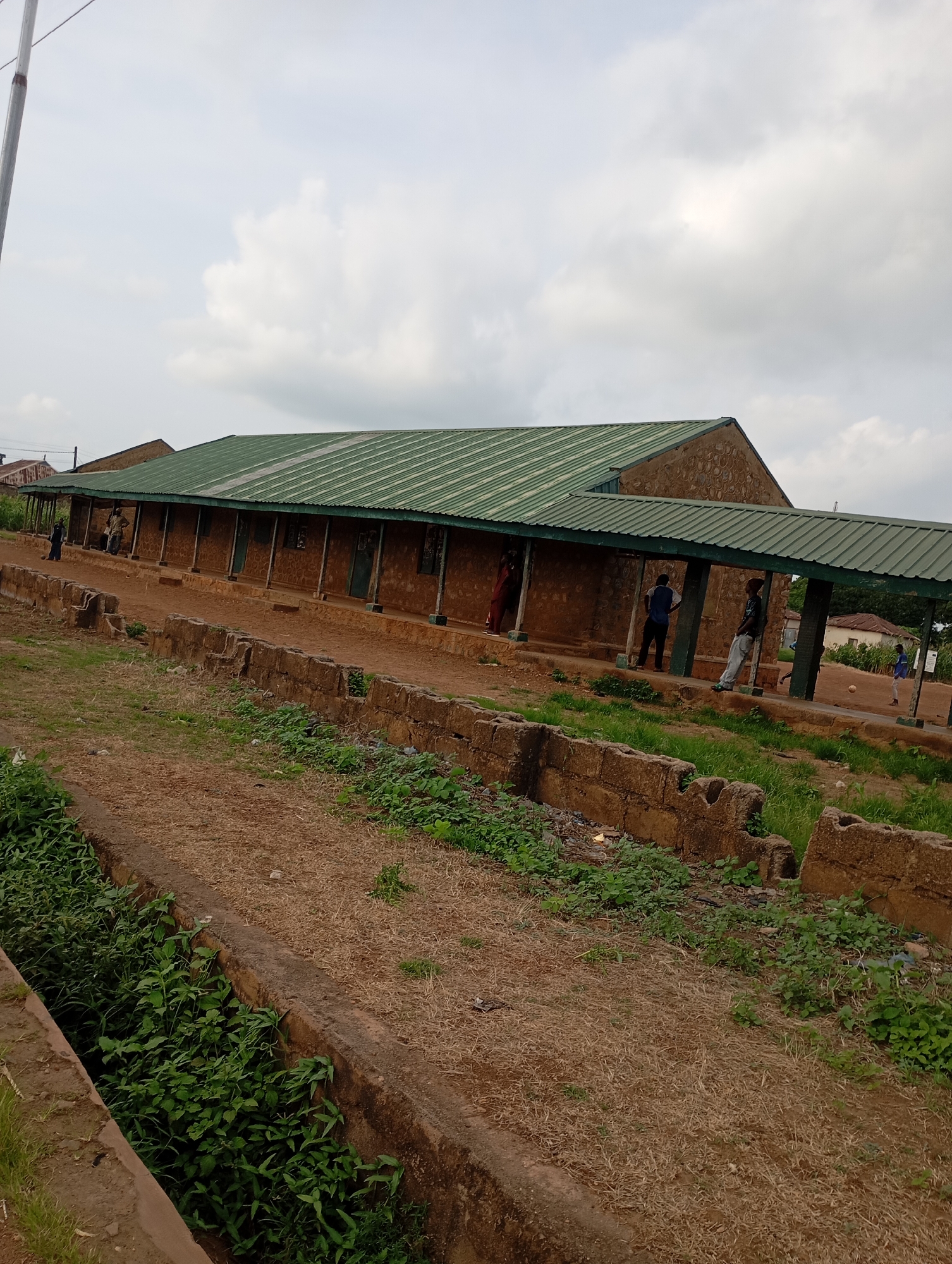The image depicts a long, single-story building with a green corrugated metal roof and a covered walkway supported by pillars. The building appears to be constructed with rough, earthy-toned blocks. Several individuals are visible in and around the building, with some standing under the walkway and others walking. The ground in front of the building is a dirt path, bordered by low, rough-hewn stone walls. Green vegetation, including grass and weeds, grows along the path and in a drainage ditch to the left of the frame. In the background, more buildings and trees are visible under a cloudy sky. The scene suggests a public facility or school in a rural or semi-urban area.