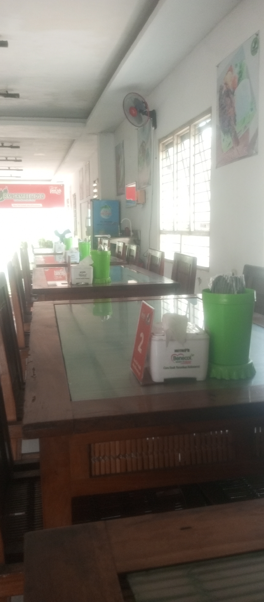 The image depicts the interior of a restaurant, likely a casual dining establishment, in Kediri City, Indonesia. The scene is dominated by wooden tables with glass tops, set up for dining. Several green plastic containers, possibly for cutlery or napkins, are visible on the tables. Some tables have white tissue boxes with branding that appears to be "Benecol". A red table marker with the number "2" is also visible on one table. In the background, there are rows of wooden chairs and what appears to be a refrigerator with a colorful label. The restaurant has a long, narrow layout, with a bright, possibly natural light source coming from windows in the background. There is a ceiling fan mounted on the wall. Posters or advertisements are displayed on the walls, including one with a red banner that reads "RM SAMBEL IDJO". The overall impression is a clean and functional dining space, characteristic of many Indonesian eateries. There are no people visible in the image.