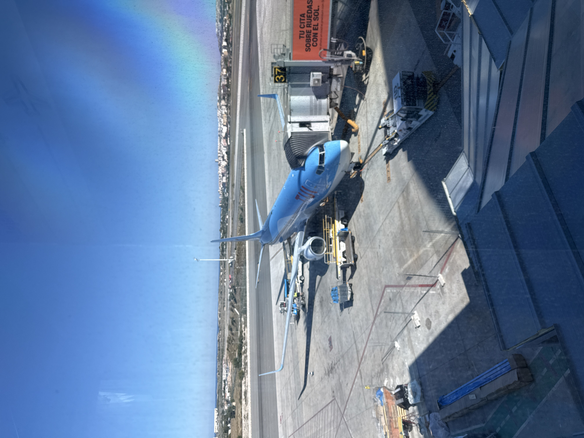 The image depicts an aerial view of an airport scene in Elx / Elche, Spain. A blue and white passenger airplane is docked at gate 37. Support equipment, including a boarding bridge and various service vehicles, surrounds the aircraft. Buildings and structures of the airport terminal and ground facilities are visible in the foreground. The urban landscape of Elx / Elche is seen in the distance, along the coastline. The sky is clear and blue with a rainbow effect in the corner of the photo, possible a reflection in the window.