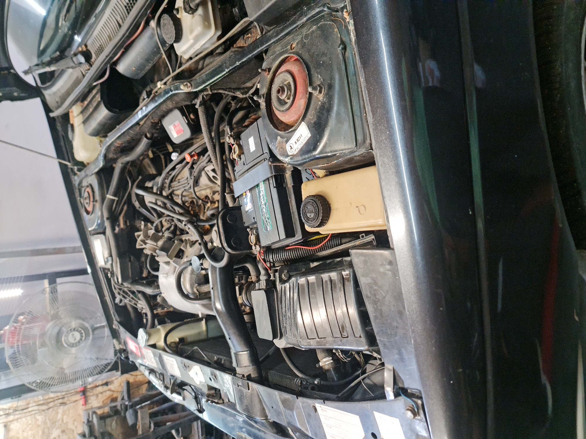 The image provides an overhead view of an open car engine bay, situated within what appears to be a garage or workshop. The scene suggests the car is undergoing maintenance or inspection.
The engine bay is filled with various components, including a black car battery prominently placed towards the right, featuring green accents and partially visible text such as "ENERGY PLUS". To its right is a beige fluid reservoir with a black cap, likely for coolant or brake fluid. A large, ribbed black air filter box is located towards the front of the engine bay. On the far right, a shock absorber's top mount is visible, characterized by a red central part secured by bolts, showing some signs of rust consistent with an older vehicle. Numerous wires, hoses, and other mechanical parts are intricately arranged throughout the engine compartment.
In the background on the left, a large, floor-standing industrial fan with a metal cage is clearly visible, suggesting ventilation or cooling in the workspace. The overall lighting appears artificial, typical of an indoor workshop environment. A faint reflection in the car's dark paintwork on the right side indicates the presence of a person standing nearby.
Visible text includes "A K 9211" on a white sticker located near the shock absorber mount. The context implies automotive repair or service, taking place in Buzovgrad, Bulgaria.