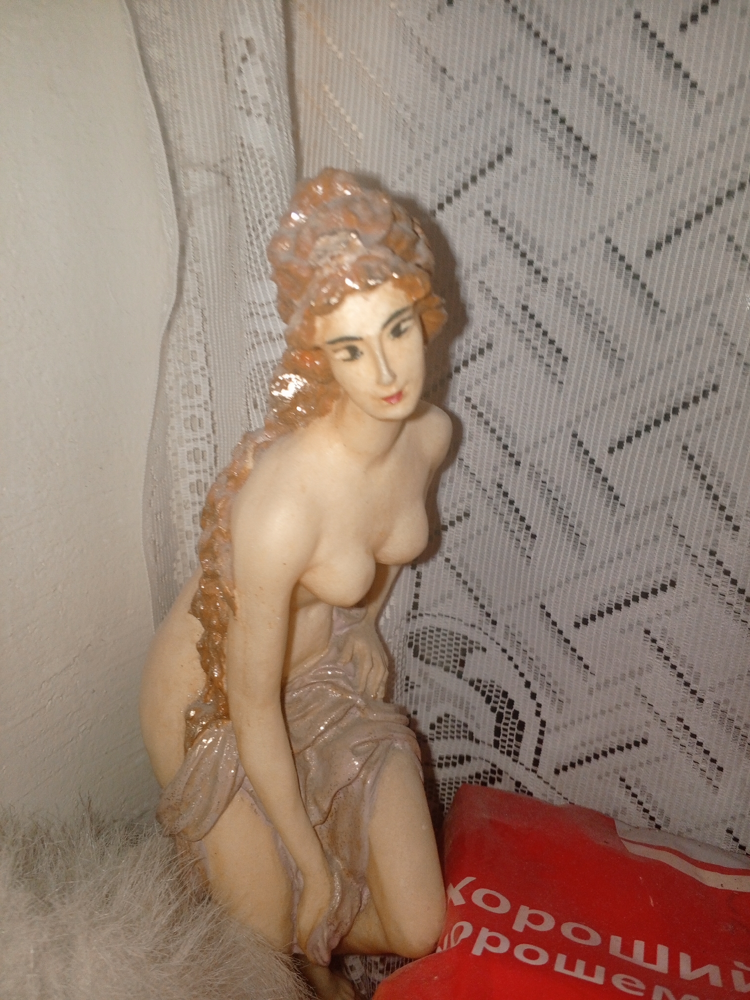 This media file features a figurine of a woman in a kneeling or reclining pose. The figurine has a light skin tone, with reddish-brown hair styled in an elaborate updo. She is adorned with a draped garment that appears to be made of a shimmering, light-colored fabric, possibly pink or beige, which is partially wrapped around her body. The figurine has detailed facial features, including painted eyes and lips. A notable detail is a golden, liquid-like substance or texture that flows down from her hair and over her shoulder and chest.
The figurine is placed in an indoor setting, with a white lace curtain or drape in the background. The texture of the curtain is intricate, with a repeating geometric pattern. To the left of the figurine, a white, textured surface, likely a wall, is visible. In the lower right corner of the image, a red object with visible text is partially cropped. The text appears to be in Cyrillic script and reads "хороший порошек," which translates to "good powder." The lighting in the scene is soft, suggesting an indoor environment, possibly illuminated by artificial light or diffused natural light. There is no indication of the time of day or weather conditions. The overall mood is still, with the figurine appearing to be a decorative item within a domestic space. The presence of the red object with text suggests it might be a product packaging, adding another layer to the observed scene.