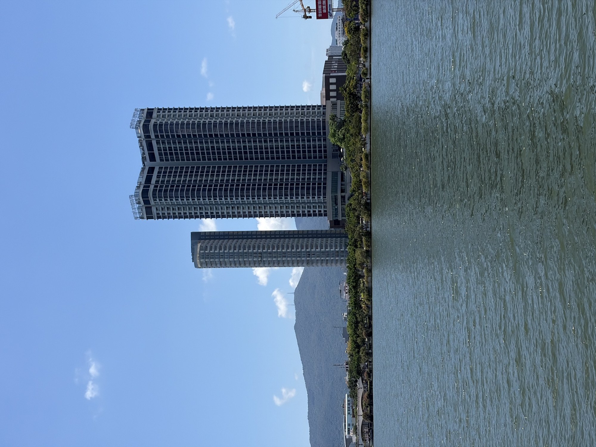 The image shows a waterfront view in Đà Nẵng, Vietnam. Two tall, modern apartment buildings dominate the left side of the frame, set against a blue sky with scattered clouds. A mountain ridge rises behind the buildings, adding to the skyline. To the right is a body of water, likely the Hàn River, extending towards the edge of the frame. Along the waterfront, a line of trees and low-rise structures is visible. A construction crane stands further in the distance.