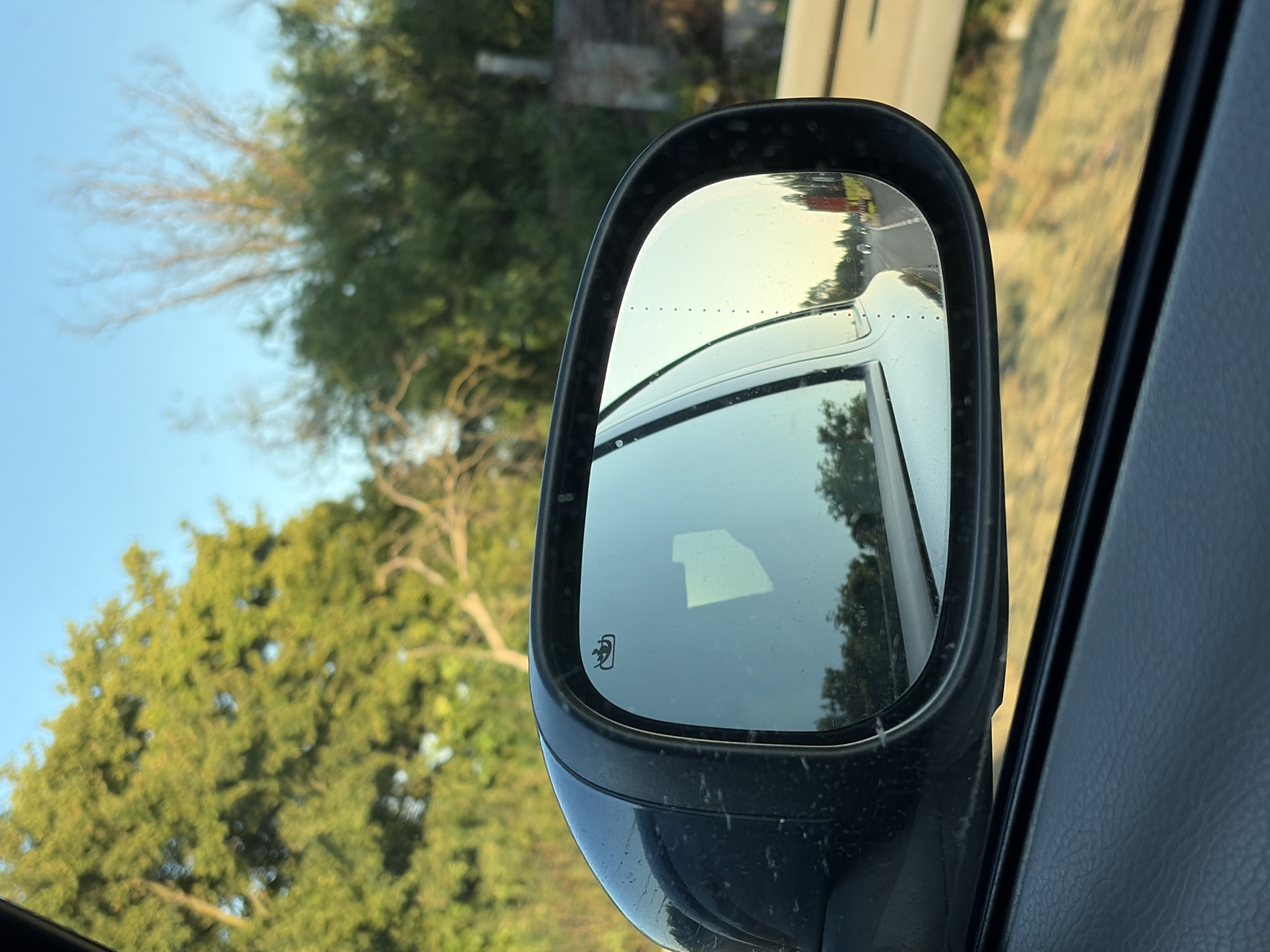 A side-view mirror of a vehicle reflects a highway scene. The mirror is black and mounted on the car door. The reflection shows a road with cars, trees, and a red truck. The sky is clear and blue. The surrounding environment is green and appears to be rural. The image is taken from inside the vehicle, looking outwards.