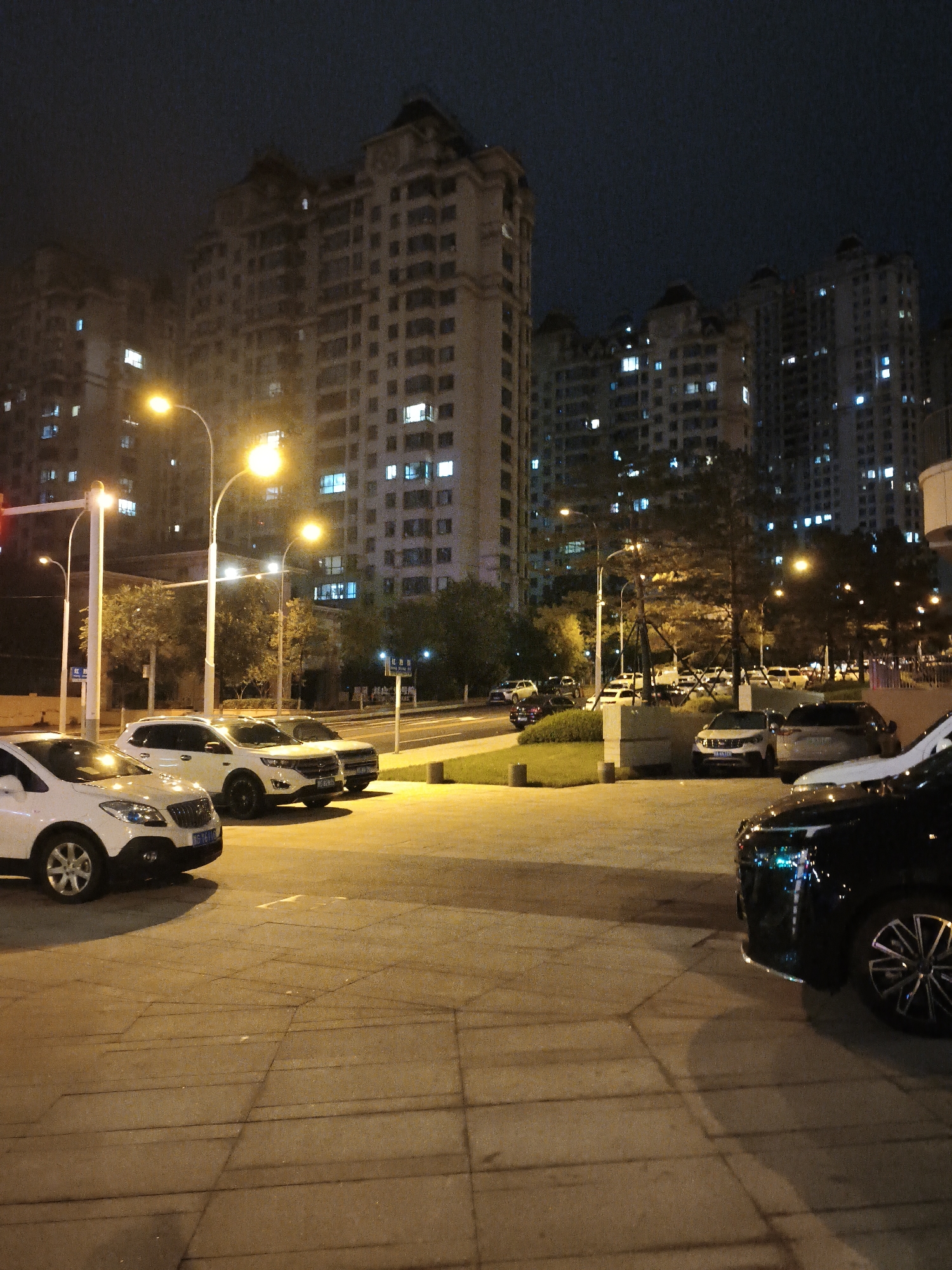 A nighttime street scene in Jixi, China, shows several parked cars on a paved area illuminated by streetlights. In the foreground, a black car is partially visible on the right. To its left, a white SUV is parked, followed by another white SUV and a white sedan. The background features tall apartment buildings with numerous illuminated windows, indicating the presence of residents. Trees are scattered throughout the scene, and streetlights cast a warm glow on the pavement. The sky is dark, characteristic of nighttime.