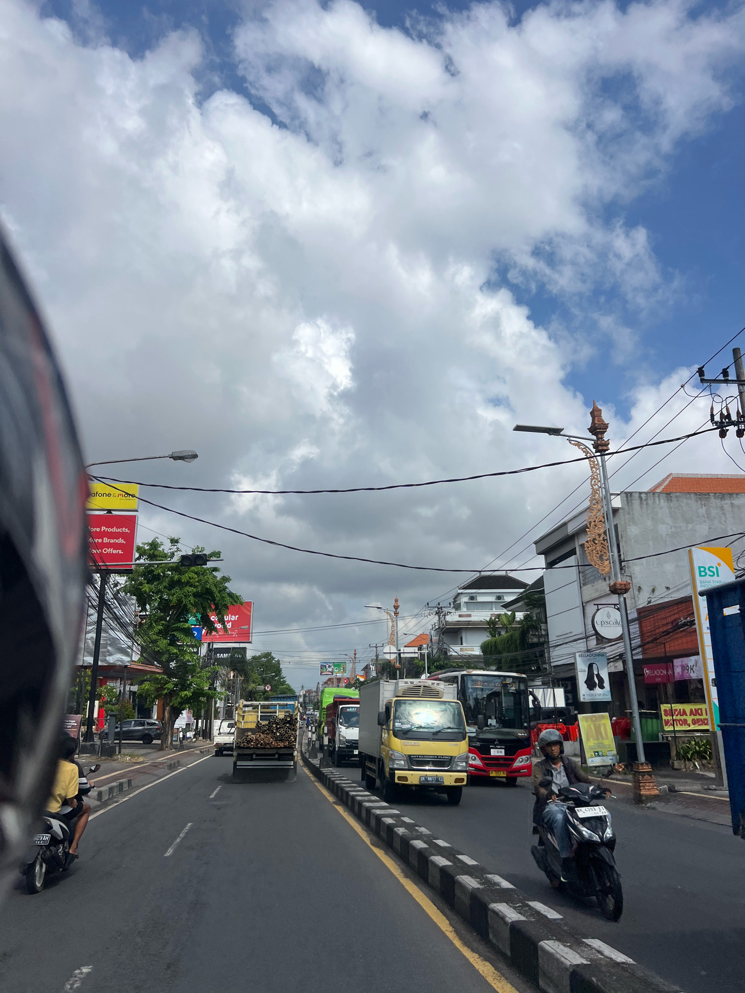 The image captures a busy street scene in Denpasar, Indonesia, likely on a daytime with a partly cloudy sky. The perspective suggests the photo is taken from the back of a motorcycle.
In the foreground, the left side of a motorcycle helmet is visible, partially obscuring the view. A person wearing a yellow t-shirt and dark shorts is riding a motorcycle on the left lane of the road.
The road itself is asphalt with yellow and white lane markings, and a concrete divider with black and white markings separates lanes. Several vehicles are on the road, including a yellow truck carrying what appears to be lumber, a green truck, and a red bus. Another motorcycle with a rider wearing a helmet and a jacket is in the right lane, moving forward.
Buildings line the right side of the street, featuring a mix of modern and older architecture. Some buildings have awnings and balconies. Several signs are visible on the buildings, including advertisements for "Vodafone & More," "BSi Bank Syariah Indonesia," and "Belladonna." Streetlights and utility poles with many wires are also present.
The environment suggests a tropical climate, with green trees visible between the buildings. The sky is blue with scattered white and gray clouds. The overall impression is one of urban activity and traffic congestion in Denpasar.