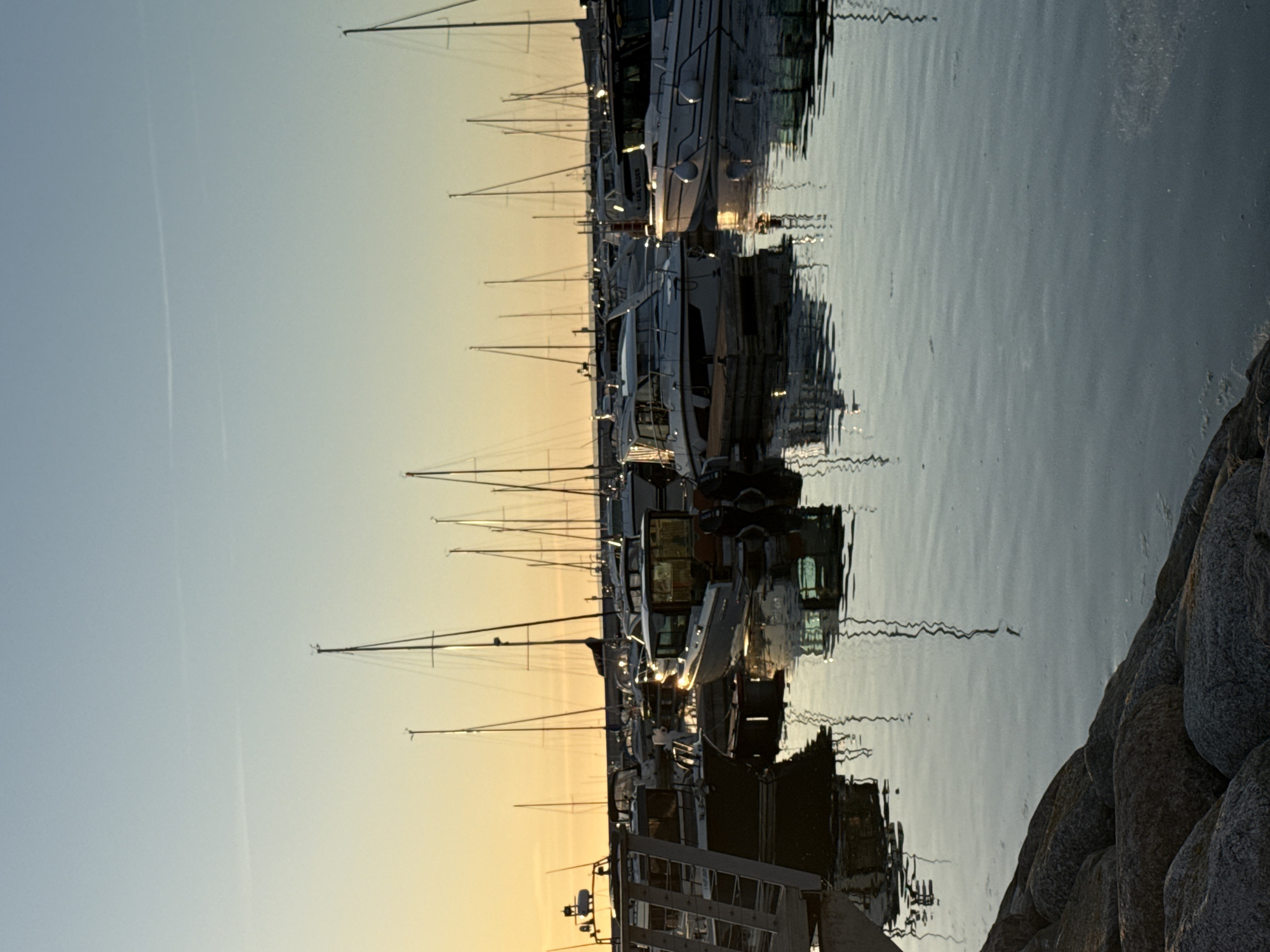 This image captures a serene view of a recreational marina in Tallinn, Estonia, during either sunrise or sunset. The low sun, positioned just outside the frame to the left, casts a warm, golden-orange hue across the horizon, gradually transitioning to a pale, clear blue in the upper sky. Two distinct, thin contrails are visible horizontally in the upper atmosphere.
The marina is densely populated with numerous boats, including both sailboats with tall, slender masts and various modern motorboats, all appearing to be docked. The boats' forms, particularly the intricate rigging of the sailboats, are reflected with notable clarity on the calm, dark surface of the water, creating a symmetrical yet slightly distorted visual. Several boat cabins and windows brightly reflect the intense golden sunlight. Within the main cabin of a prominent light-colored motorboat towards the center-left, two dark, indistinct figures are faintly discernible as silhouettes.
The water, which occupies the majority of the lower half of the image, is remarkably still with subtle ripples, accurately mirroring the docked vessels and the gradient of the sky. The immediate foreground on the bottom-left reveals a dark, rugged rocky shoreline, partially submerged in the water. The backlighting from the low sun creates strong silhouettes of the boats and their masts against the bright horizon, emphasizing the tranquil atmosphere of the harbor.