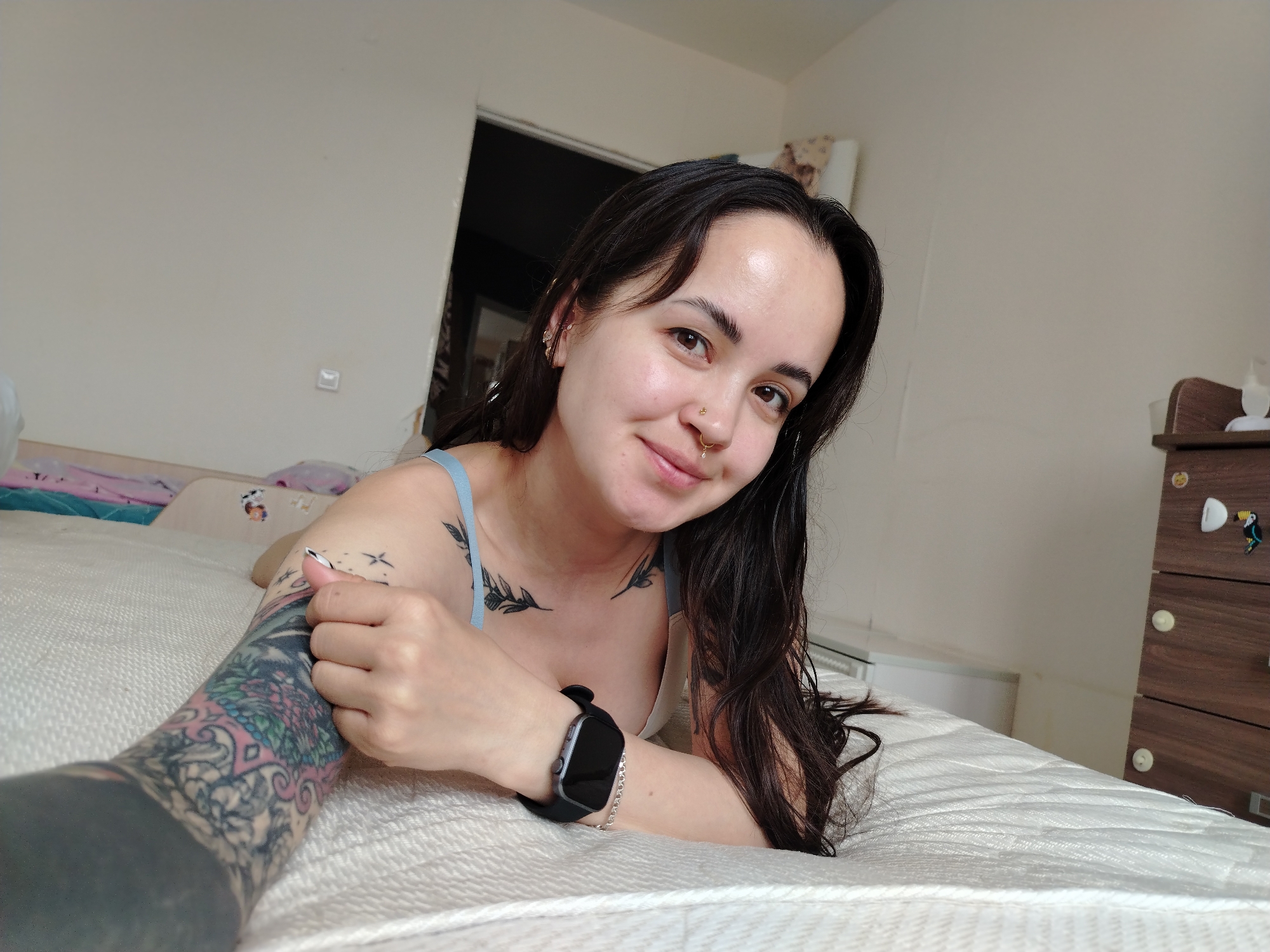 A woman lies on a bed, smiling directly at the camera. She has dark hair, a nose ring, and several tattoos visible on her arms and chest. She is wearing a wristwatch on her left wrist. The bed is covered in a white mattress pad. Behind her is a white wall with a doorway leading to a dark room. To the right, a wooden dresser is visible. The location is not evident from the image. There is no information suggesting that the photo was taken in Noviy, Russia.