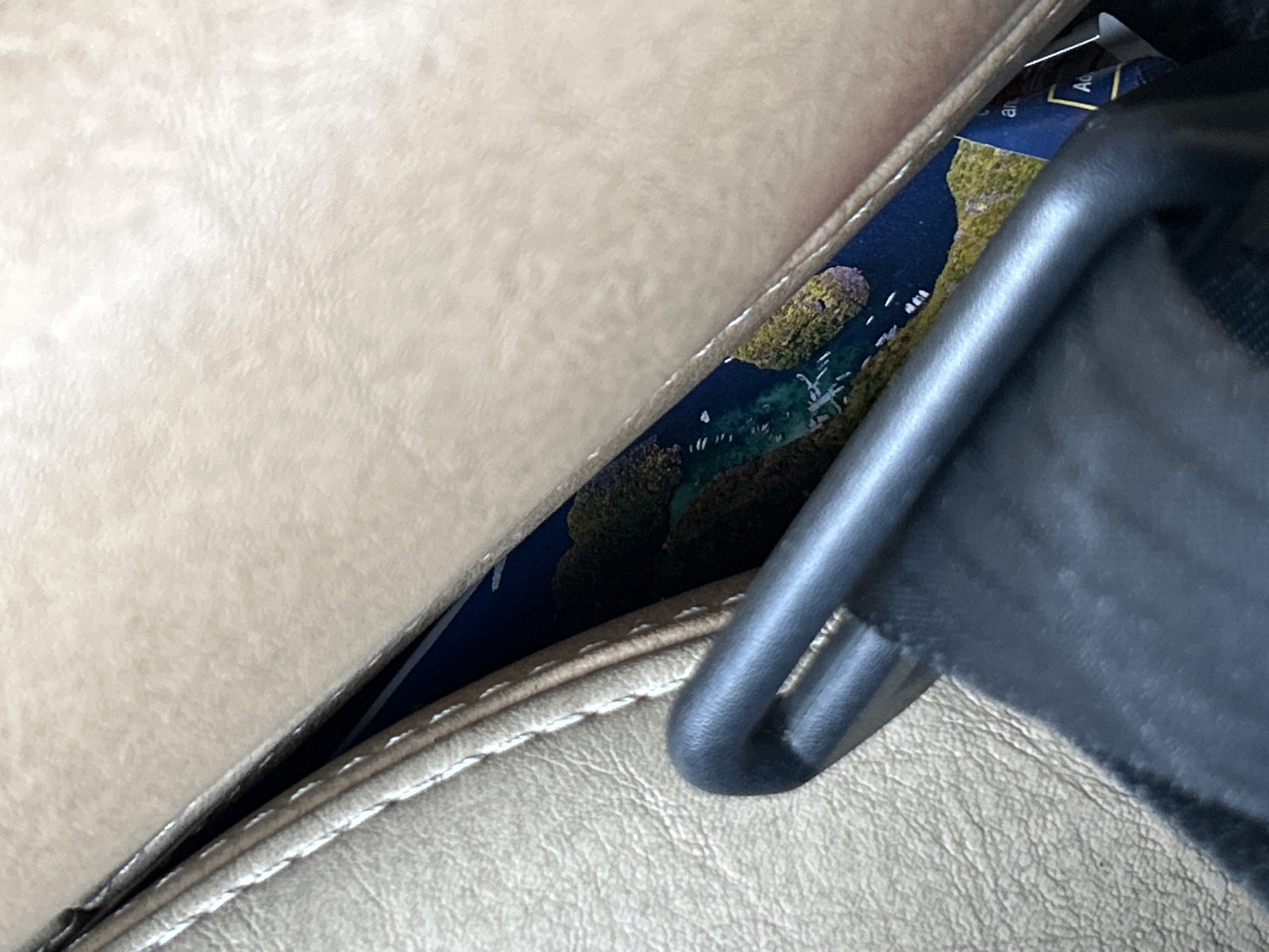 The image shows a close-up view of a beige leather seat, possibly a car or airplane seat, with a seatbelt buckle visible in the foreground. Tucked into a crevice of the seat is a small rectangular object, appearing to be a postcard or travel brochure. The visible portion of the object features a dark blue background with an aerial image of a tropical landscape, showing green islands with bodies of water. Text is partially visible on the object, with the letters "an" and "Ad" discernible. The overall scene suggests a travel-related context, and the presence of the seatbelt implies transportation. Without further context, the specific location in Phuket City Municipality, Thailand cannot be determined from the image alone.