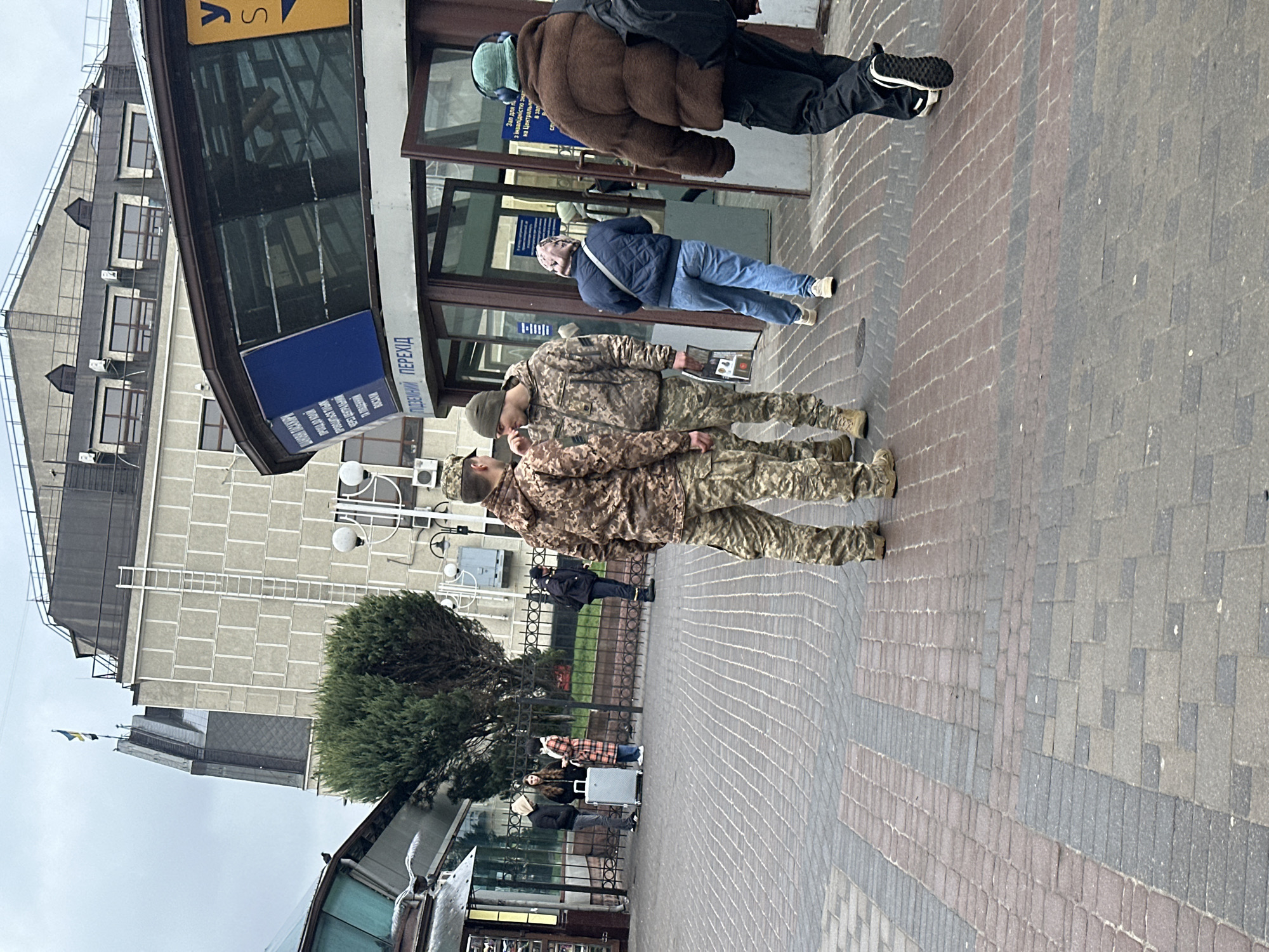 The image depicts a street scene in Kyiv, Ukraine. Two soldiers in camouflage uniforms stand on a brick sidewalk, appearing to be in close conversation. Behind them are pedestrians passing by, including a person in a brown puffer jacket and another in a blue jacket. In the background, there is a building with a sign that reads "ПІДЗЕМНИЙ, ПЕРЕХІД," indicating an underground pedestrian crossing. The building also features signs in Ukrainian. A small Ukrainian flag is visible atop another building in the distance. Additional people are present in the background, some near a fenced area. The street is paved with alternating shades of brick.