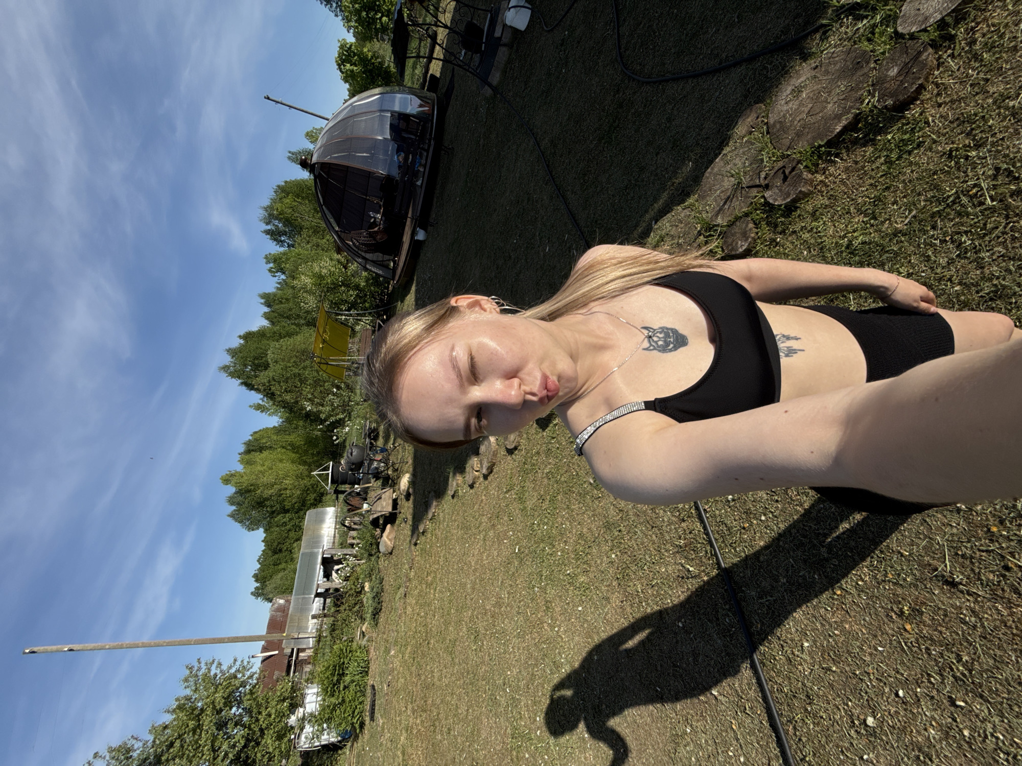 A blonde woman in a black top and shorts is taking a selfie outdoors. The setting appears to be a rural area in Unknown, Russia, with a house, trees, and a dome-shaped structure visible in the background. The grass is green and sunlit. The woman is wearing jewelry and has tattoos on her chest.