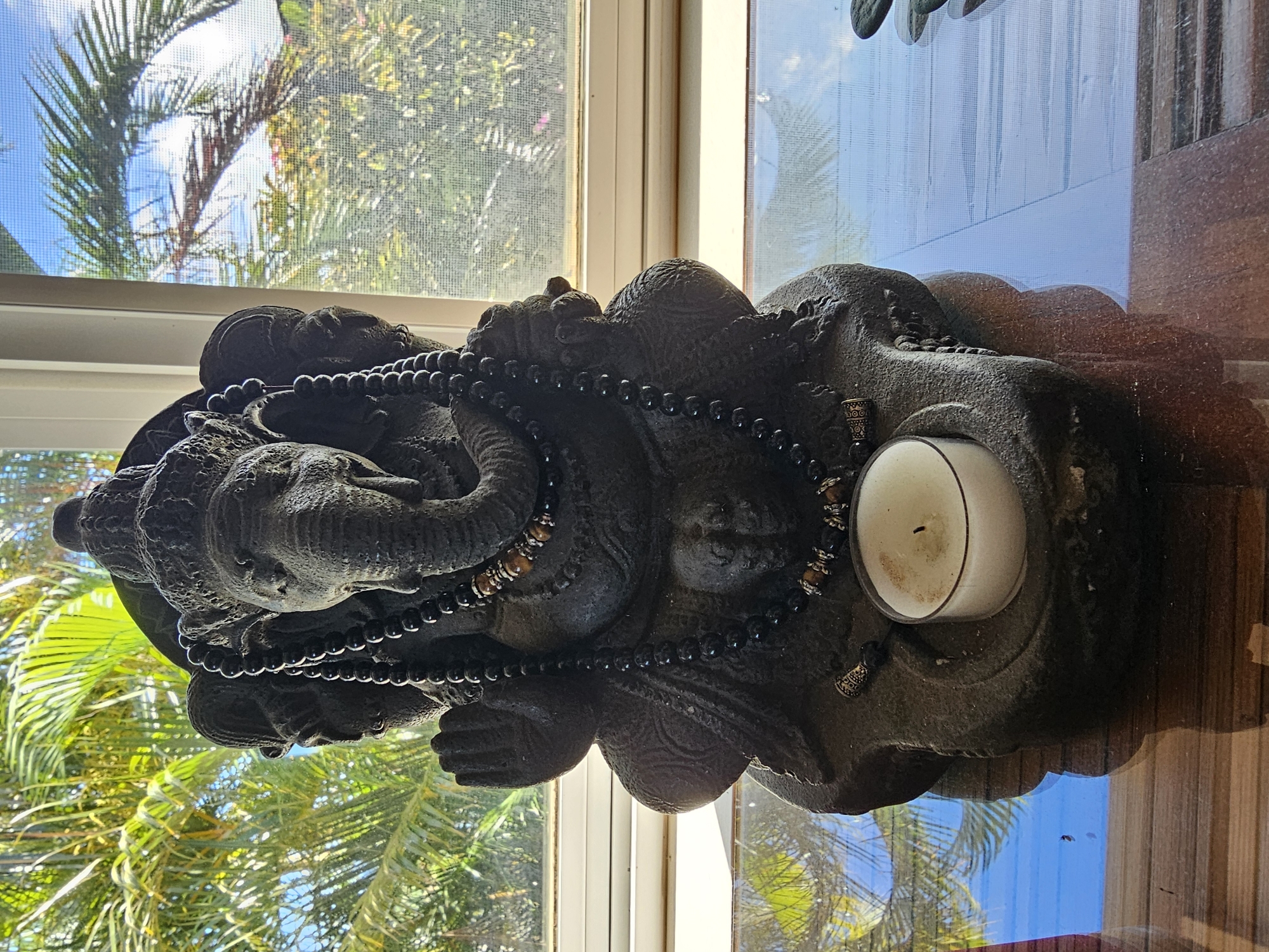 An indoor close-up shot depicts a dark stone statue of the Hindu deity Ganesha seated with his hands in a mudra, adorned with black beaded necklaces. The statue is placed on a wooden surface in front of a window with sheer white blinds, through which palm trees and a bright blue sky are visible. A lit white tea candle sits in a stone holder at the base of the statue. The setting is likely Napili, United States of America, given the tropical foliage outside the window.