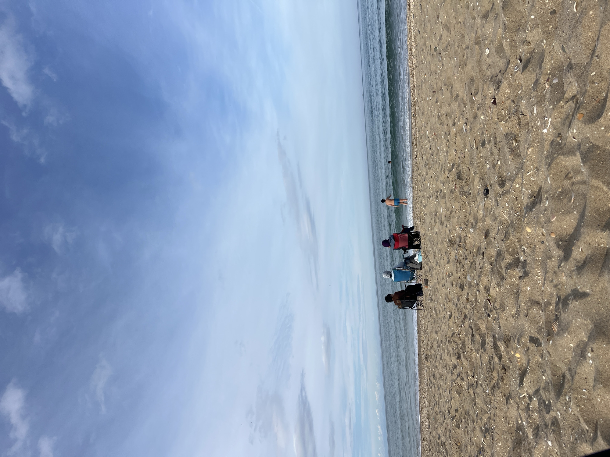 The image depicts a beach in M'Diq, Morocco, on a clear day with a blue sky and scattered clouds. The beach extends along the right side of the frame, covered in sand and seashells. Three beach chairs are positioned near the water's edge, occupied by people. A man is standing in the water, further down the shoreline. The ocean stretches out towards the horizon.