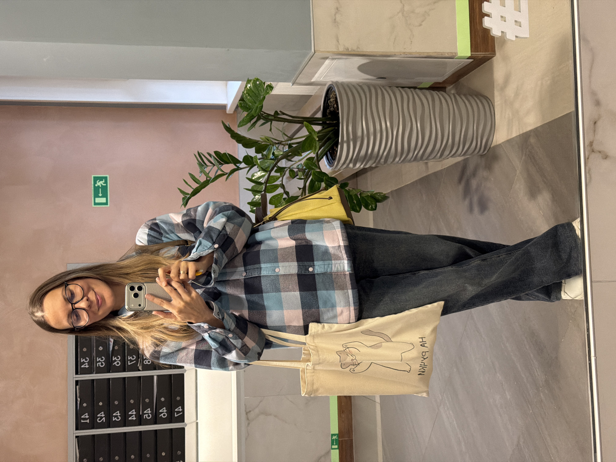 This is a vertical, eye-level, indoor shot of a young woman taking a mirror selfie. She is wearing a blue and pink plaid collared shirt, dark wash jeans, and white sneakers. She has blonde hair, glasses, and is smiling at the camera. She is holding a silver smartphone in her right hand, and a yellow clutch bag is tucked under her left arm. A canvas tote bag with a line drawing of a cat and some text is hanging from her shoulder.
In the background, to her left, are a row of black mailboxes, some with numbers visible. Above the mailboxes is a pale pink wall. To her right, a large potted plant with dark green leaves is placed in a decorative, grey, ribbed planter. Further to the right, a section of wall displays a white hashtag symbol. The floor is tiled with light grey and beige marble-look tiles. The lighting appears to be artificial, suggesting it is daytime or evening. The scene suggests a residential building hallway or lobby. There is a green "exit" sign with a running figure symbol on the pink wall. The text on the tote bag is in Cyrillic script.