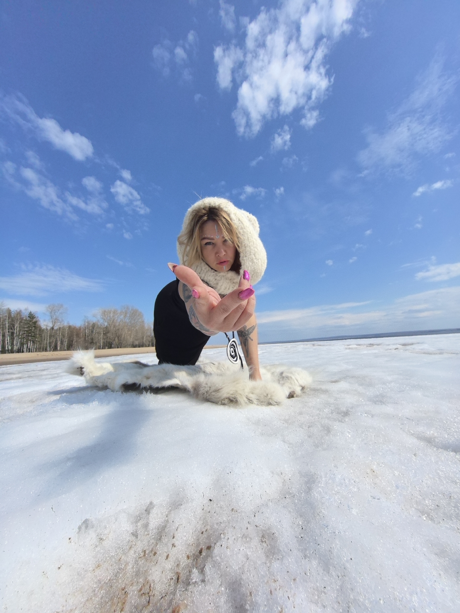 A wide-angle, low-angle shot captures a woman posing in a snowy, outdoor landscape under a bright blue sky with scattered white clouds. The woman, with light hair, is positioned low to the ground, appearing to kneel or recline on snow and ice. She wears a cream-colored, knitted hooded garment, a dark top, and a distinctive black and white circular pendant necklace. Light markings or makeup are visible on her face, above her eyebrows and on her chin. Her right hand, adorned with vibrant pink nail polish and tattoos, is extended towards the viewer with an open palm. A white or light-colored fur-like garment is spread beneath her on the icy surface.
The background features a distant treeline of bare deciduous trees, beyond which lies a vast body of water, likely a lake or sea, hinting at a large expanse of frozen or partially frozen water. The foreground shows glistening snow and ice, with some patches appearing to be partially melted or slushy, reflecting the bright sunlight. The time of day is clearly bright daylight, suggesting a sunny, cold, and clear day. The woman's direct gaze and pose create a sense of engagement with the camera. The setting aligns with a cold, northern environment, consistent with the provided location context of Noviy, Russia. There is no visible text in the image.