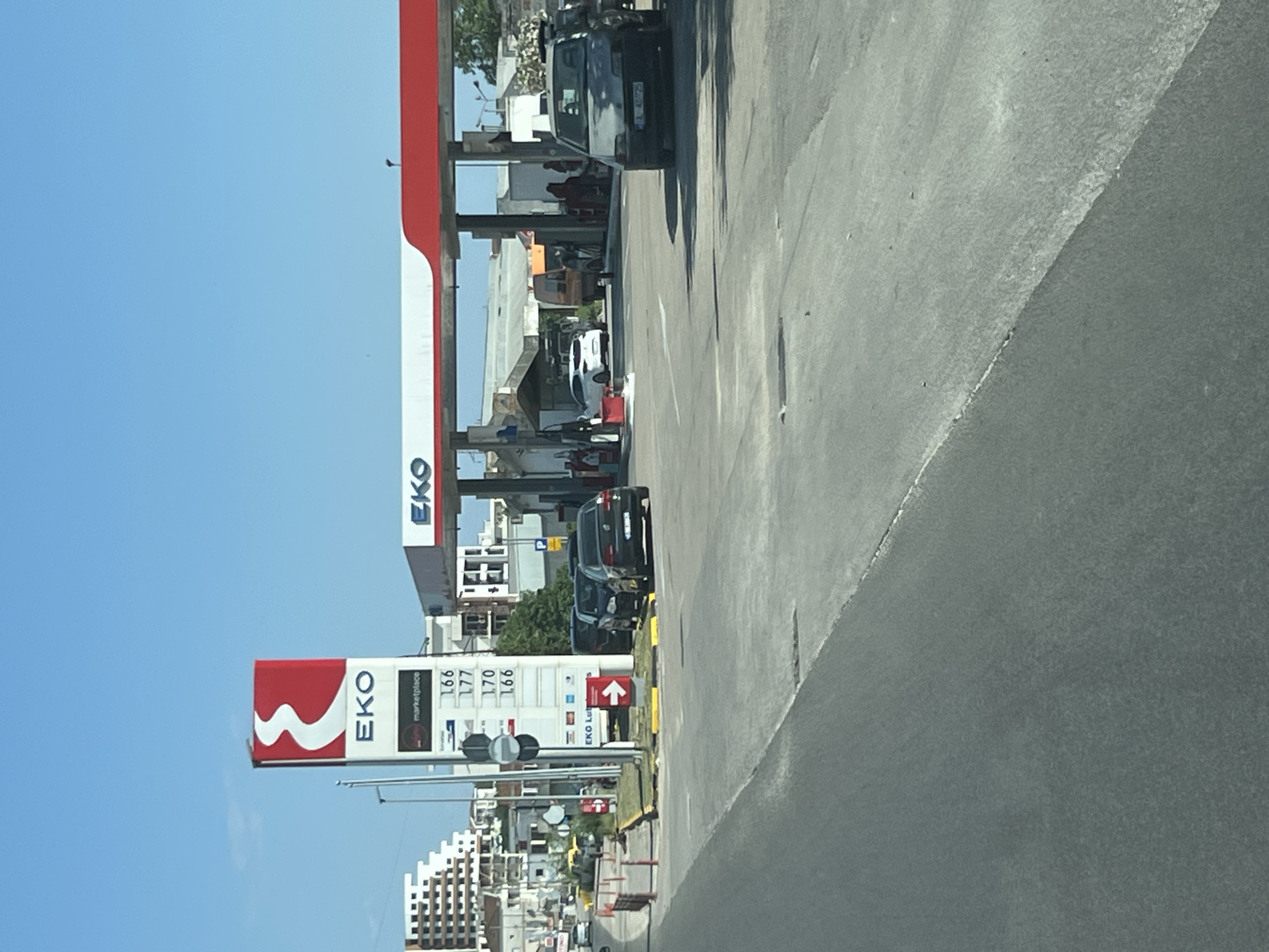 This media file captures a sunny daytime scene at an EKO gas station in Ulcinj - Ulqin, Montenegro. The perspective is from a vehicle, looking towards the gas station and the road.
The primary subjects are the gas station infrastructure, including the prominent red and white EKO branding on the canopy and a tall sign displaying fuel prices. Several cars are visible, some are at the pumps, suggesting the activity of refueling. One dark-colored SUV is parked prominently near the pumps. People are visible under the gas station canopy, appearing to be interacting or waiting.
In the background, there are buildings, suggesting an urban or semi-urban environment. Trees are also present, adding to the outdoor setting. The sky is a clear, bright blue, indicating fair weather. The road surface is visible in the foreground, appearing somewhat worn and textured.
Notable details include the fuel prices displayed on the EKO sign, with numbers such as "166," "177," and "170" visible. The EKO logo, a stylized red "W," is clearly identifiable. The overall impression is of a functional, active gas station on a pleasant day.