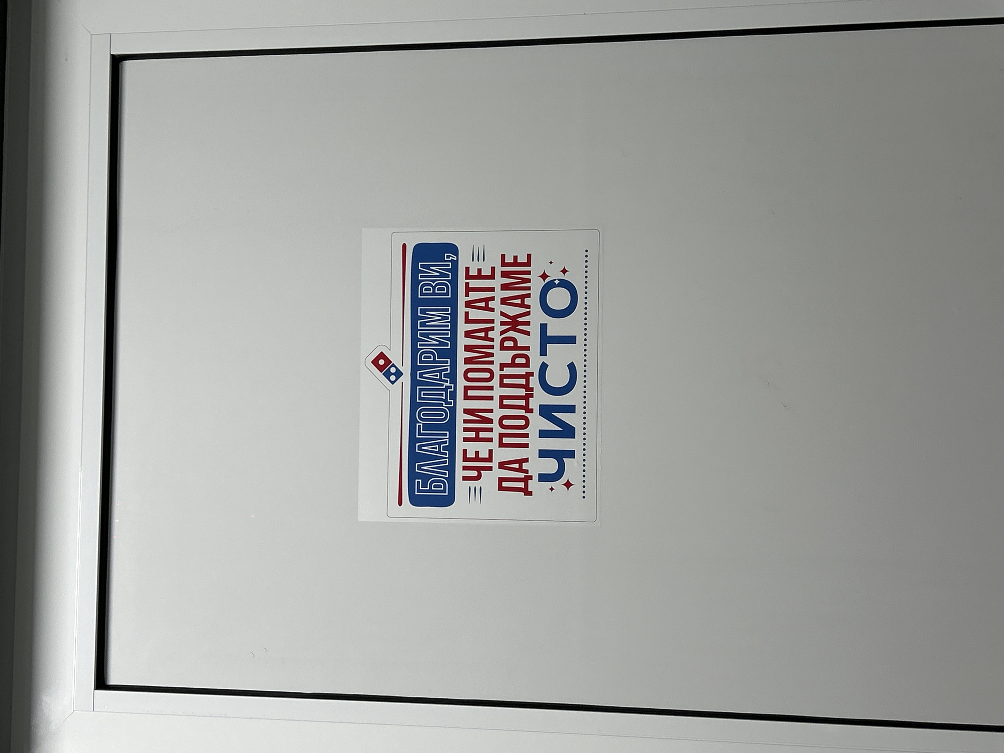 A close-up, eye-level shot captures a clean, white door or panel, likely part of an interior space within a building in Burgas, Bulgaria. The door features a simple, modern design with a thin black line marking the edge of the panel and a white frame. Centrally placed on the door is a rectangular sticker, primarily white, adorned with a message in Bulgarian and the Domino's Pizza logo.
The sticker's design uses bold blue text for the first line and red text for the subsequent lines, with the word "ЧИСТО" (CLEAN) highlighted and accompanied by small red sparkle graphics. The Domino's Pizza logo, displaying its iconic red and blue domino tile design, is positioned to the left of the Bulgarian text.
The text on the sticker reads:
"БЛАГОДАРИМ ВИ," (THANK YOU,)
"ЧЕ НИ ПОМАГАТЕ" (FOR HELPING US)
"ДА ПОДДЪРЖАМЕ" (TO MAINTAIN)
"ЧИСТО" (CLEAN)
The message is an expression of gratitude for assistance in maintaining cleanliness. The overall setting appears well-maintained and orderly, consistent with a commercial establishment, likely a Domino's Pizza restaurant, emphasizing hygiene. No people or activities are visible, focusing solely on the door and its message.