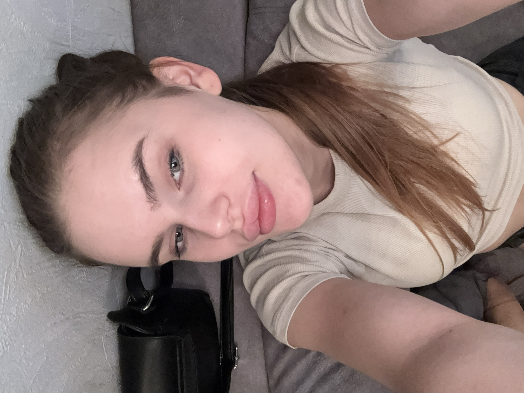 A young woman is captured in a selfie, lying down on a couch with a grey textured wall behind her. Her head is slightly tilted, and she is looking towards the camera with a soft smile. She has light brown hair pulled back into a ponytail, and her eyes are a striking blue-grey. She is wearing a light-colored, ribbed, short-sleeved shirt. A black purse with a silver buckle is visible next to her. The lighting suggests it is daytime. The scene appears to be indoors, likely in a home or living space in Dnipro, Ukraine. The overall mood is relaxed and casual. There is no visible text in the image.