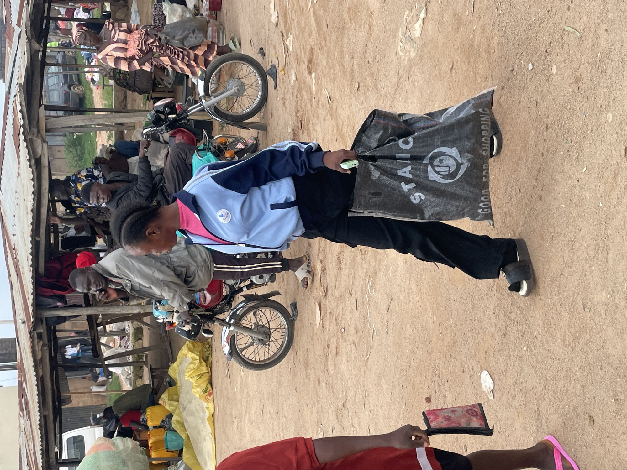 In Wang, Nigeria, a market scene unfolds with several individuals and objects. A young woman in the foreground, wearing a blue and white jacket and black pants, carries a large black shopping bag. She walks from left to right across a dusty, unpaved ground. Behind her, a motorcycle is parked. In the background, a covered market stall is visible, with various goods and people interacting. One man, on his phone, stands near a motorcycle, while another person sits on a bench. The market appears to be a bustling open-air environment with temporary structures.