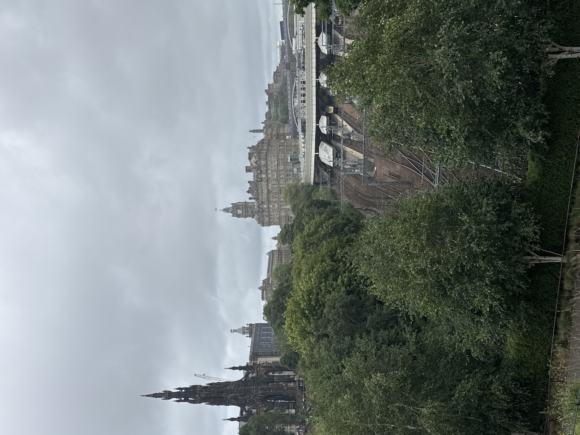 The image depicts an overcast view of a cityscape in Edinburgh, United Kingdom. In the foreground, lush green trees and bushes are visible, with a metal fence and dry grass in the immediate bottom. Behind the foliage, a complex network of railway tracks and platforms is situated, with several trains at the station. Further in the background, several notable buildings and structures are present. To the left, a tall, dark, Gothic-style monument with intricate detailing stands prominently, with a construction crane visible behind it. Adjacent to this monument are other historical buildings. In the center and to the right, a large, multi-story stone building with a clock tower dominates the skyline, appearing to be a significant landmark. Other buildings and bridges are scattered across the horizon, suggesting a dense urban environment. The sky is uniformly grey and cloudy, contributing to a muted color palette of the scene. No people are discernible in the image.