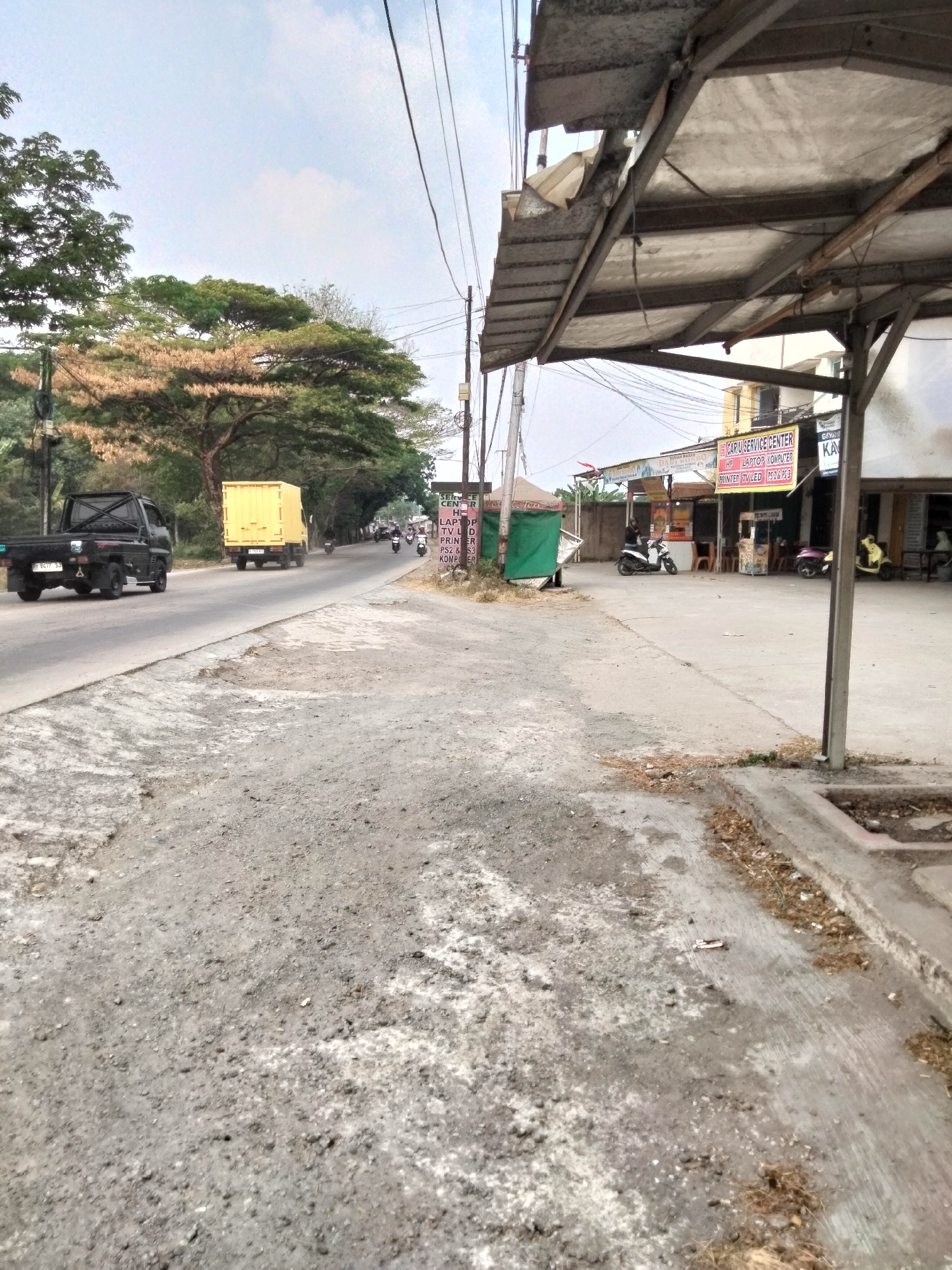 The image depicts a street scene in Cariu, Indonesia. In the foreground, a rough, gray asphalt road with scattered debris is visible. To the right, a weathered gray canopy structure with a metal frame and torn fabric covering is prominent. Beyond the canopy, a sidewalk area is seen, with a building facade featuring several shop windows and signs. One sign reads "CARU SERVICE CENTER HP LAPTOP KOMPUTER PRINTER TV LED PS2 & PS3 KOMPUTER." Various shops are open with some seating outside.
The street itself is active with traffic. A black pickup truck with license plate "B 9272 SJ" is moving away from the camera on the left side of the road. Further down the road, a yellow box truck is also traveling in the same direction. Several motorcycles are visible on the road, with riders. In the background, lush green trees line the roadside, with some trees exhibiting autumn foliage. The sky is overcast with some patches of blue. The overall impression is a commercial area along a busy road in a semi-urban setting.