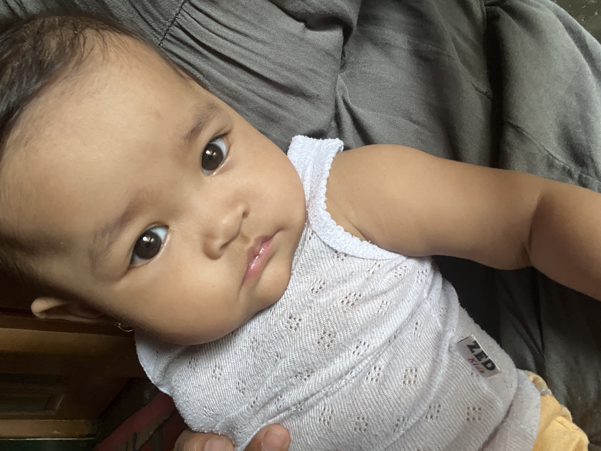 The image shows a close-up of a baby with dark brown eyes, fair skin, and dark hair. The baby is wearing a white, textured sleeveless top with a lace trim. A small gold earring is visible in the baby's left ear. The baby's face is turned slightly towards the camera, with a neutral expression. The background consists of gray fabric, likely clothing, and a dark wooden surface, possibly furniture, on the left. The baby appears to be held by someone, as a hand is visible on the left side of the image, near the baby's shoulder. A small tag with the text "ZED Kids" is attached to the baby's top. The scene is an indoor setting. The exact location in Kruengmate, Indonesia cannot be determined from the image alone.