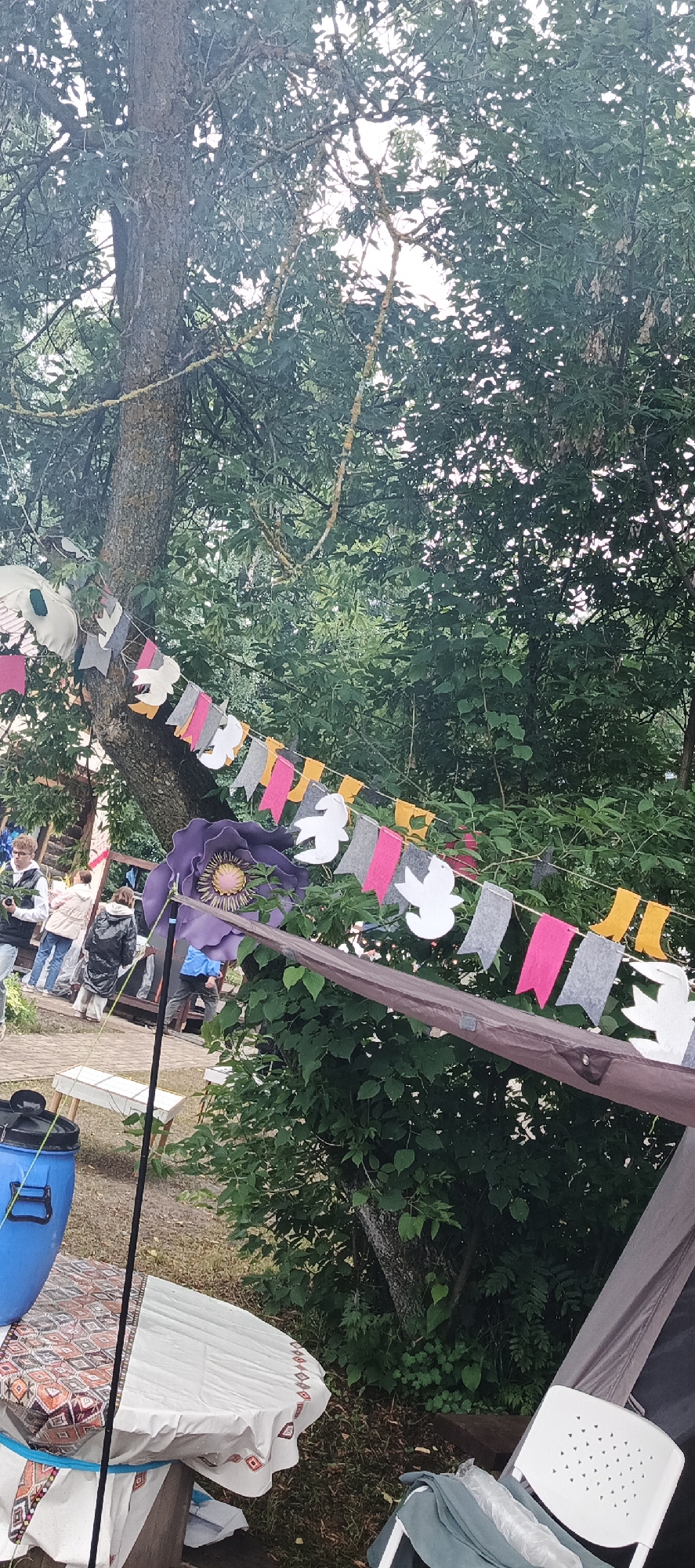 The image depicts an outdoor scene, likely a park or recreational area, with lush green trees forming a canopy. A string of colorful bunting, featuring flags in pink, grey, yellow, and white cutouts of birds or doves, is strung between trees. In the foreground, a large purple flower decoration is prominently displayed, attached to a pole. To the left, a blue plastic bin sits on a table covered with a white and patterned cloth. Several people are visible in the background, engaged in various activities, with some appearing to be walking or standing around. The overall atmosphere suggests a casual outdoor gathering or event. The location is identified as Старые Быги, Russia.