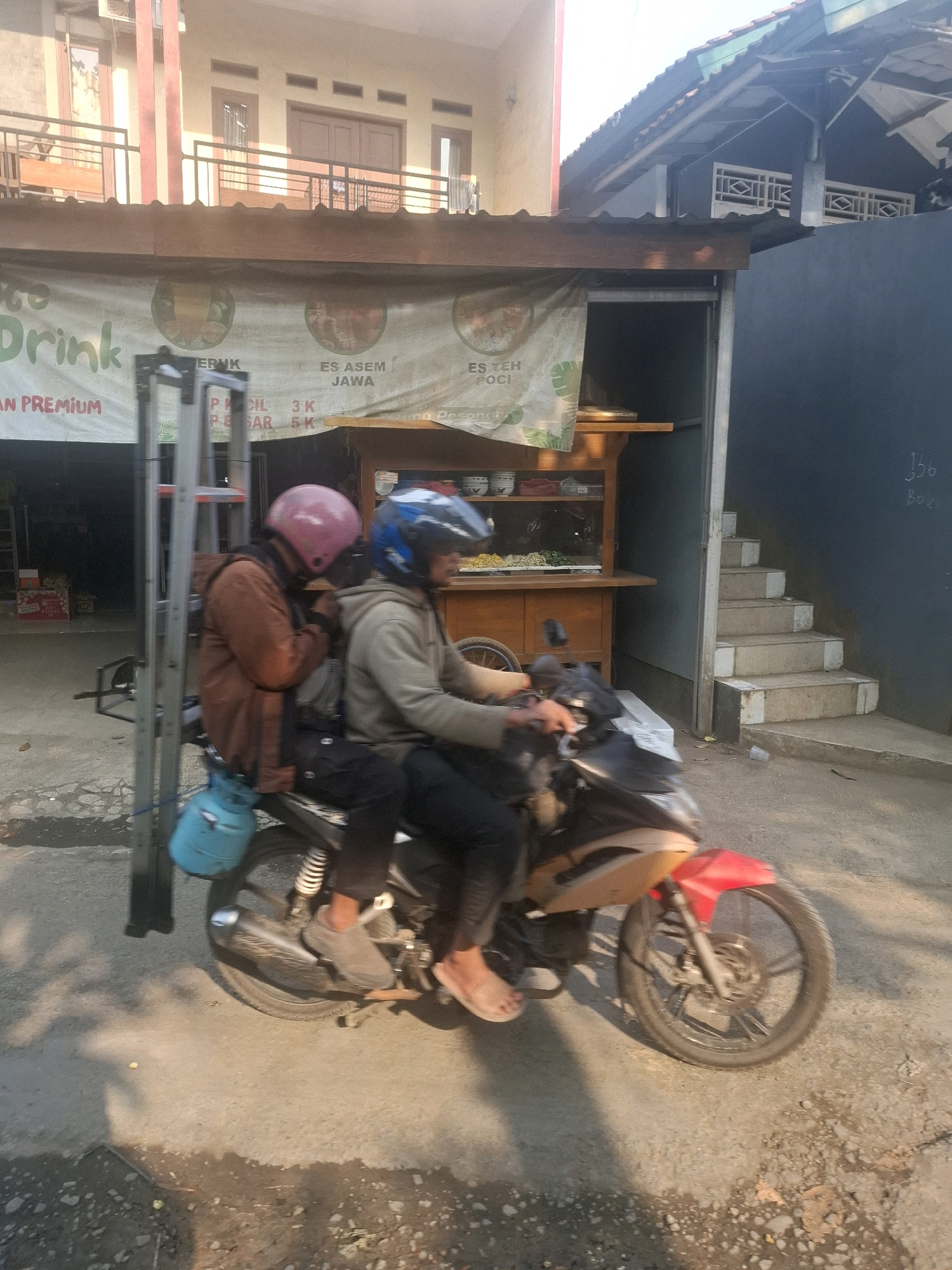 Two individuals are riding on a motorcycle in what appears to be a street in Ciheulang Tonggoh, Indonesia. The person in front is wearing a pink helmet and a brown jacket, while the person behind them is wearing a blue helmet and a grey hoodie. Both are wearing dark pants. A light blue gas tank is attached to the back of the motorcycle. A metal ladder is leaning against the side of the motorcycle. In the background, there is a building with a banner advertising drinks, a wooden food stall with various items displayed, and a set of concrete stairs leading up to a dark blue wall. The scene is illuminated by sunlight, casting shadows on the ground.