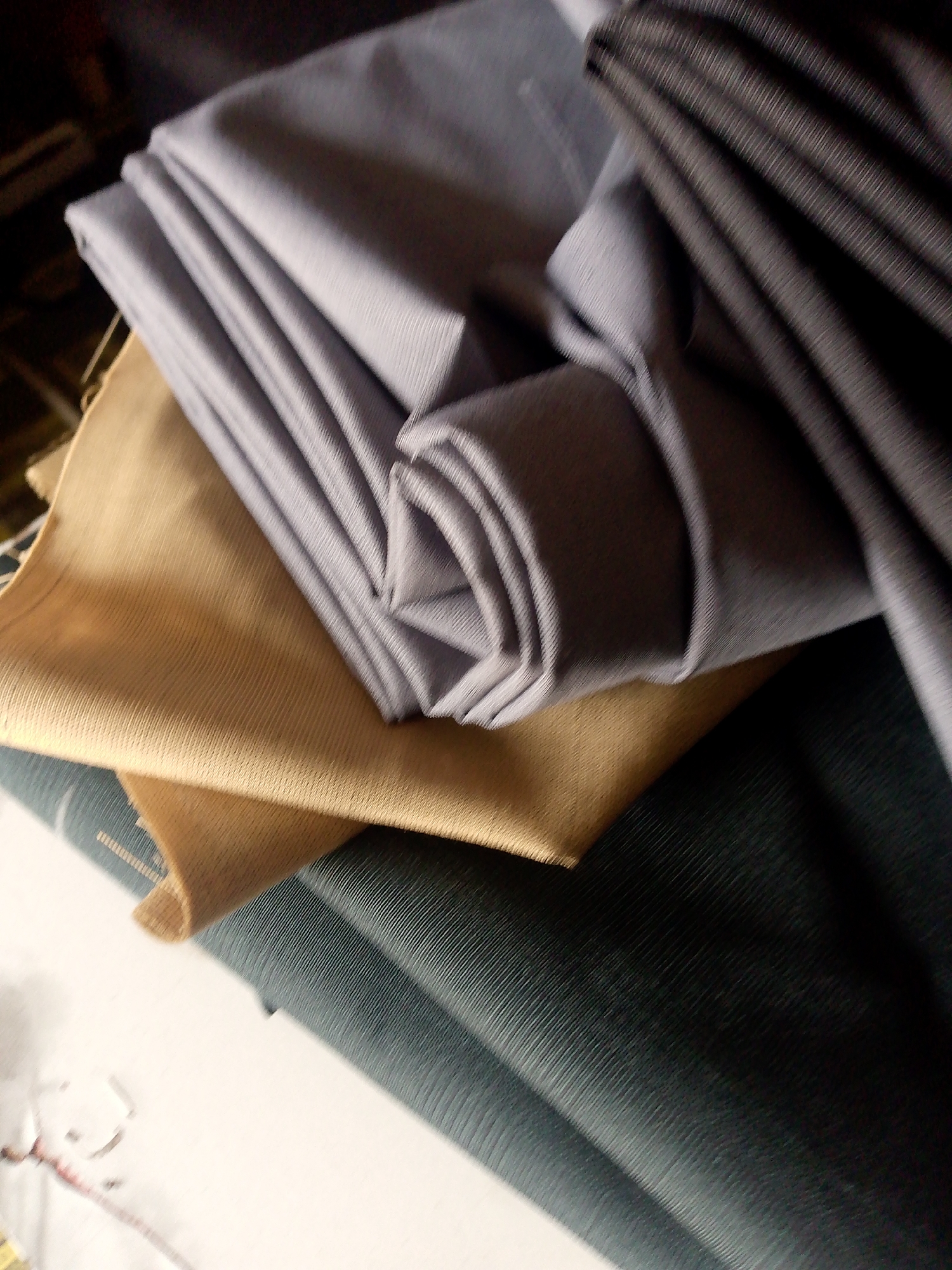 In the city of Jauro Yinu, Nigeria, a close-up shot captures a stack of folded fabrics. The top layer is a light grey material with fine vertical ribbing, folded into a bundle. Beneath it lies a tan fabric, appearing to be a coarser weave. A dark grey fabric with prominent vertical ridges is visible on the right, draped over the other materials. The bottom fabric is a dark teal color, also with a ribbed texture. The lighting suggests an indoor setting, possibly a shop or storage area.