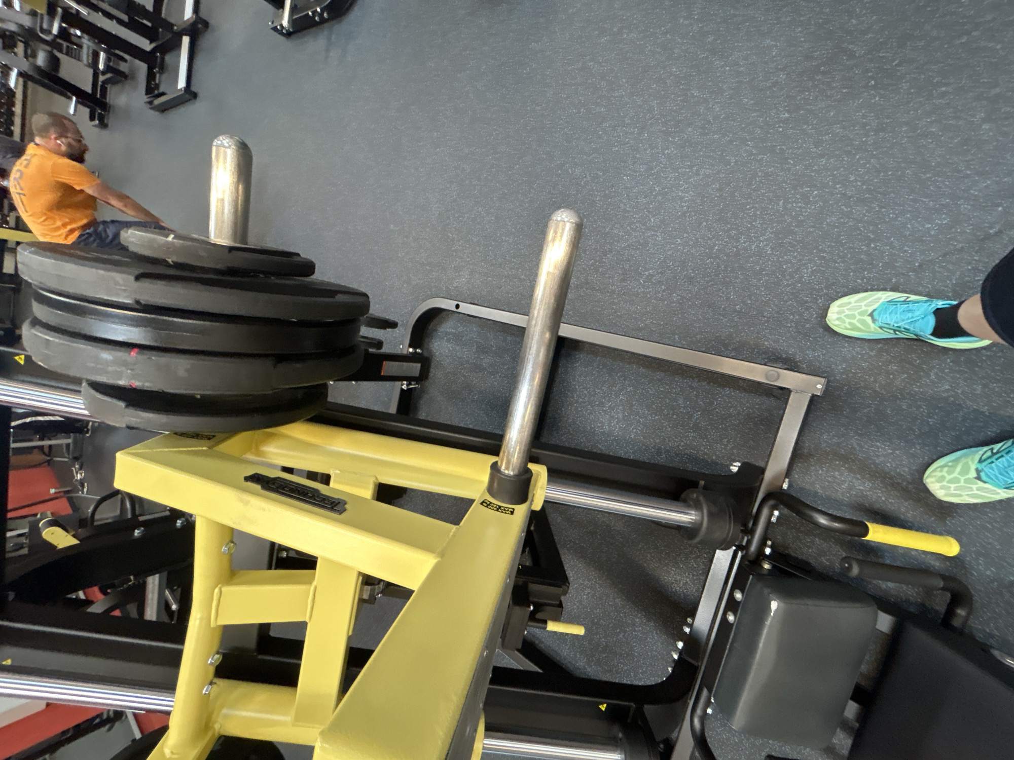 The image presents an overhead view of gym equipment in a gym environment, likely located in Sofia, Bulgaria. A yellow and black leg press machine is in the foreground, loaded with several weight plates on a silver post. To the right of the machine are two feet wearing green and blue patterned running shoes. In the background, a man wearing an orange shirt appears to be using a separate piece of gym equipment. The gym floor is dark gray. Other gym equipment is partially visible in the periphery, providing context to the location. The image focuses on the leg press machine, the weight plates, and the feet of a person presumably using the machine.