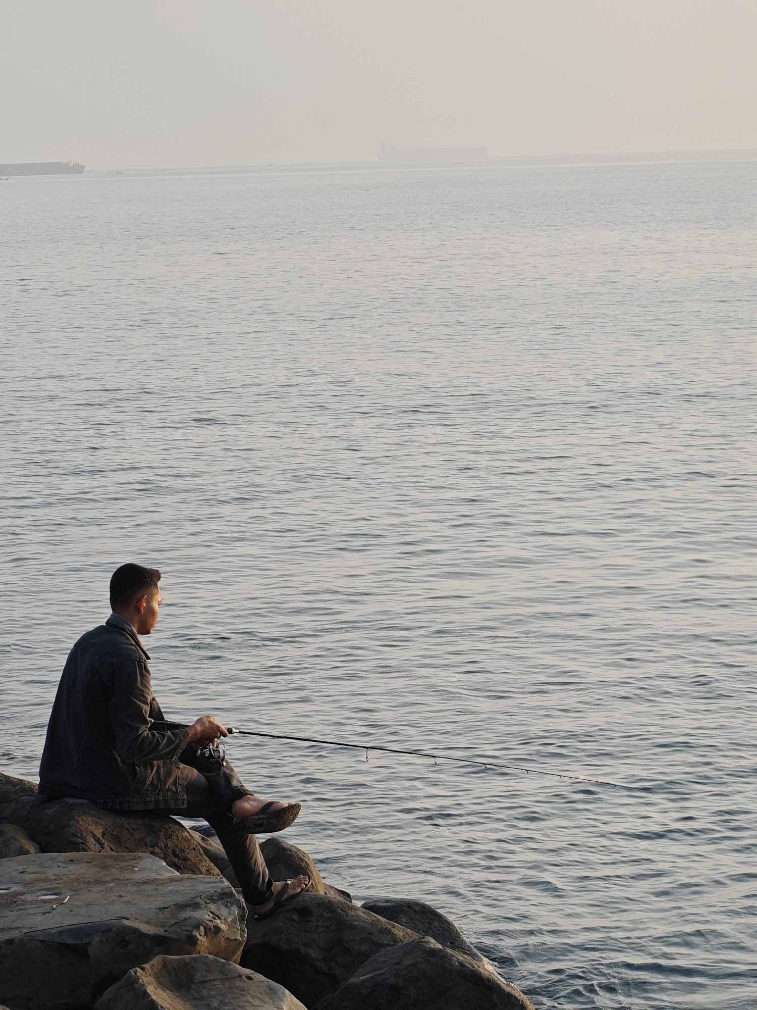 A person sits on large rocks by the ocean in Lhokseumawe, Indonesia, holding a fishing rod. The person, wearing a dark denim jacket and pants, is facing away from the camera, looking out at the water. The sea is calm with small ripples, and the sky is overcast, creating a muted color palette. In the distance, a large cargo ship is visible on the horizon. The rocks are rough and uneven, suggesting a coastal area. The overall atmosphere is serene and solitary.