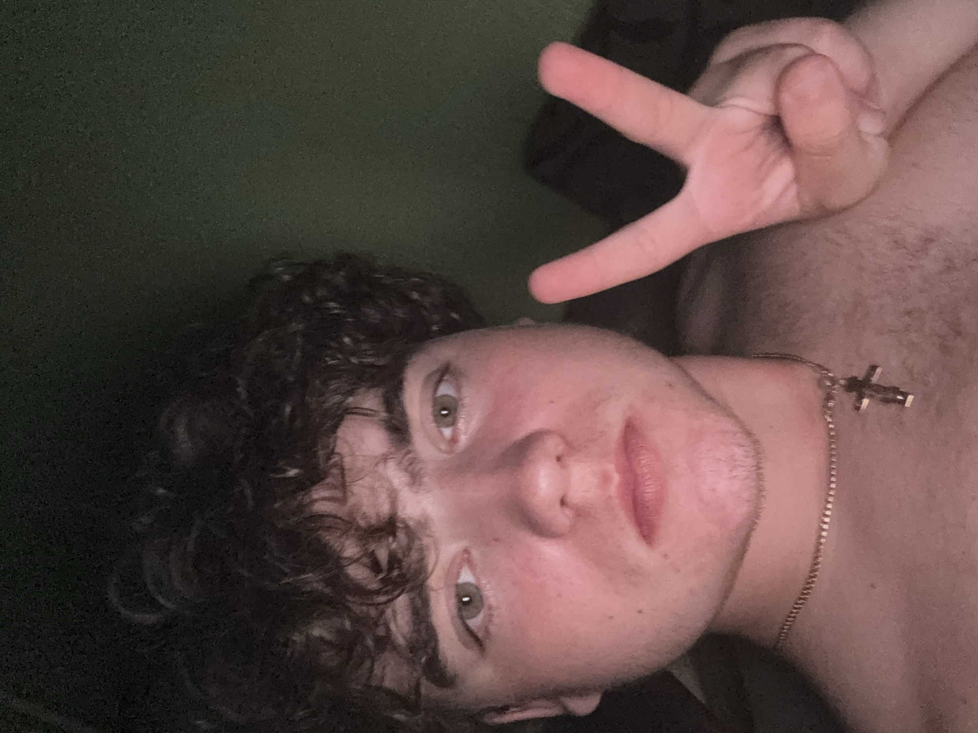 The image is a selfie of a young man with curly brown hair. He is looking directly at the camera and holding up his right hand in a peace sign. He is wearing a gold chain with a cross pendant. The background is dark green and appears to be a wall. The lighting is dim, suggesting the photo was taken indoors at night. The image does not contain any information that could indicate its location in Zaporizhzhia, Ukraine.