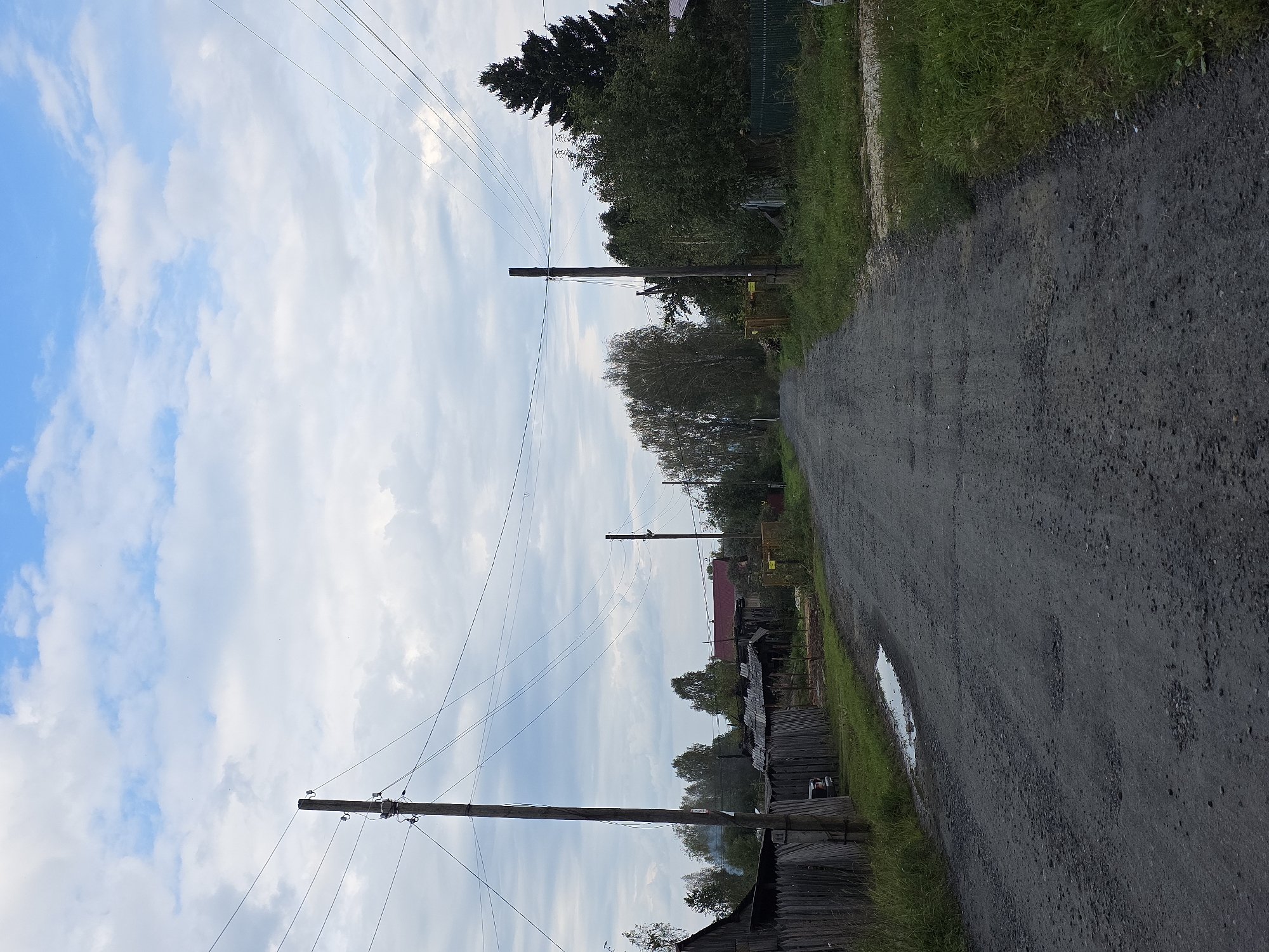 The image depicts a view of a dirt road in the city of Magnitka, Russia, under a partly cloudy sky. The road is in disrepair, with numerous potholes visible. On the left side of the road, a wooden fence runs along a property with a dilapidated structure behind it. A utility pole with wires stands prominently near the fence. Further down the road, more trees and structures can be seen, including a house with a red roof. On the right side of the road, dense greenery and another utility pole with wires are present. The scene suggests a rural or semi-rural setting within Magnitka.