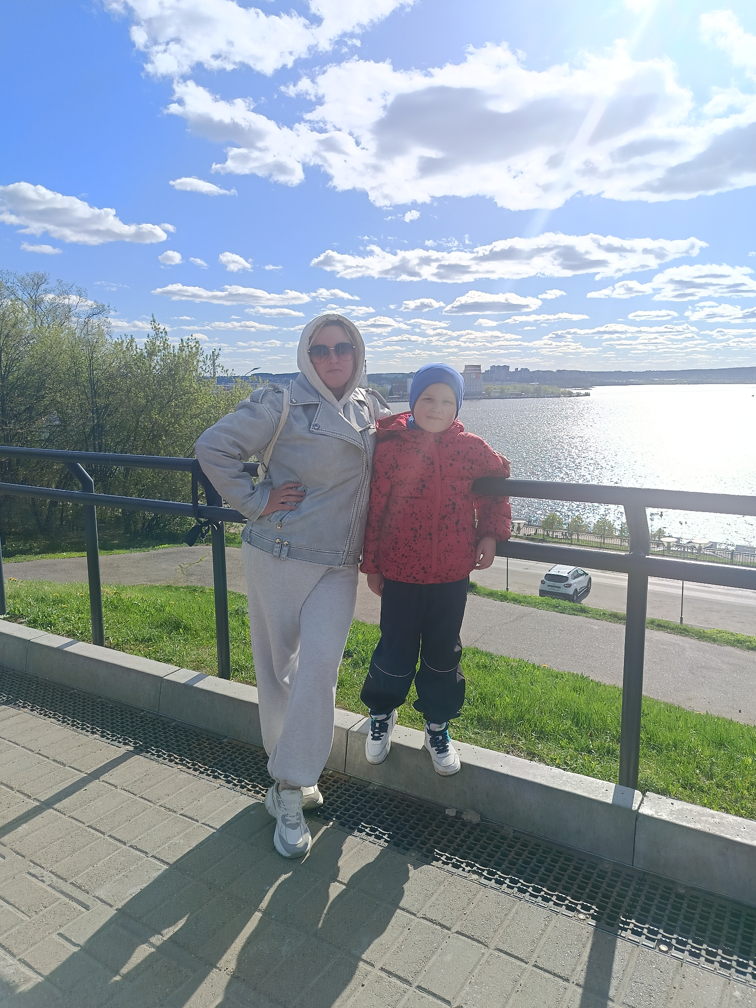 The image shows a woman and a boy standing in front of a railing overlooking a body of water.
The woman is wearing a light-colored hooded jacket, light-colored sweatpants, and white sneakers. She is wearing sunglasses and has her hand on her hip.
The boy is wearing a red jacket, black pants with light-colored cuffs, and a blue hat. He is standing on the ledge of a concrete barrier, leaning against the railing.
Behind the railing is a body of water, and on the opposite shore, there are buildings, indicating an urban setting. A car is visible on a road below.
The sky is blue with scattered white clouds, and the sun is shining brightly.
Based on visual cues like the architecture, the body of water, and the overall environment, the location appears to be in Izhevsk, Russia. The scene likely depicts a park or scenic overlook in the city.