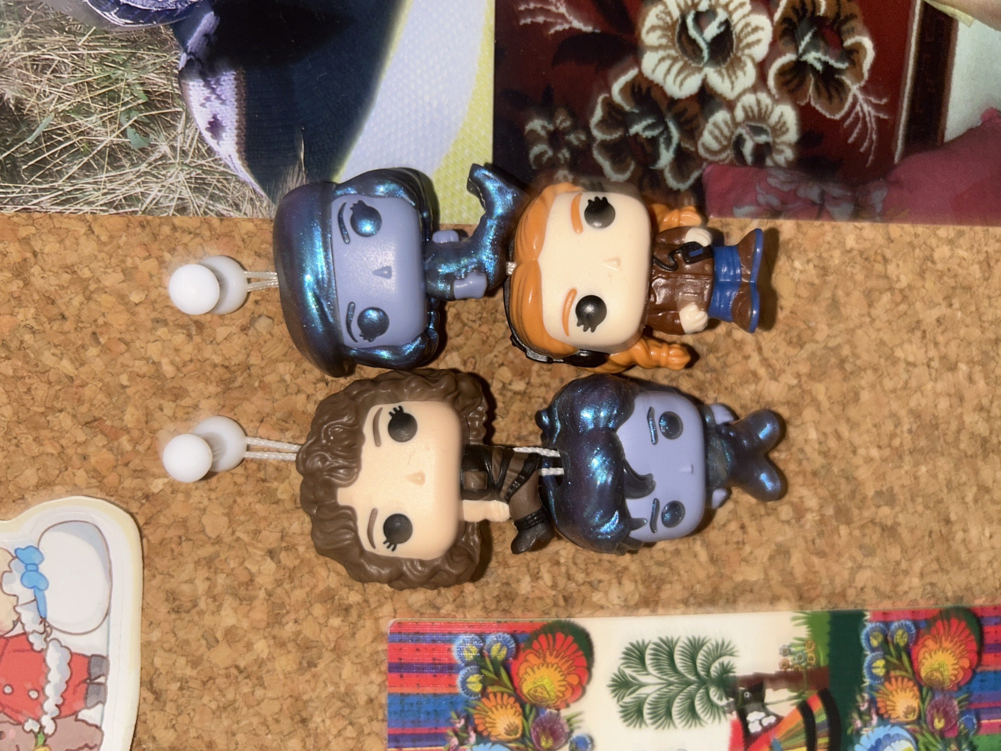 The image shows a corkboard with several items pinned to it. There are four Funko Pop figures of characters with oversized heads and simplified features pinned to the board. There are two blue figures, one brown-haired figure, and one red-haired figure. They appear to be attached with straight pins with round white heads. In the upper left corner is a portion of a photo showing grass and a dark-colored object. In the upper right corner is a portion of a patterned fabric or wallpaper with floral motifs. In the bottom left corner is a small section of a colorful cartoon-style image with a scalloped edge. In the bottom right corner is a fragment of a postcard or similar item with a vibrant floral design. The background is a plain cork surface. The image does not provide information regarding the city of Câmara de Lobos, Portugal.