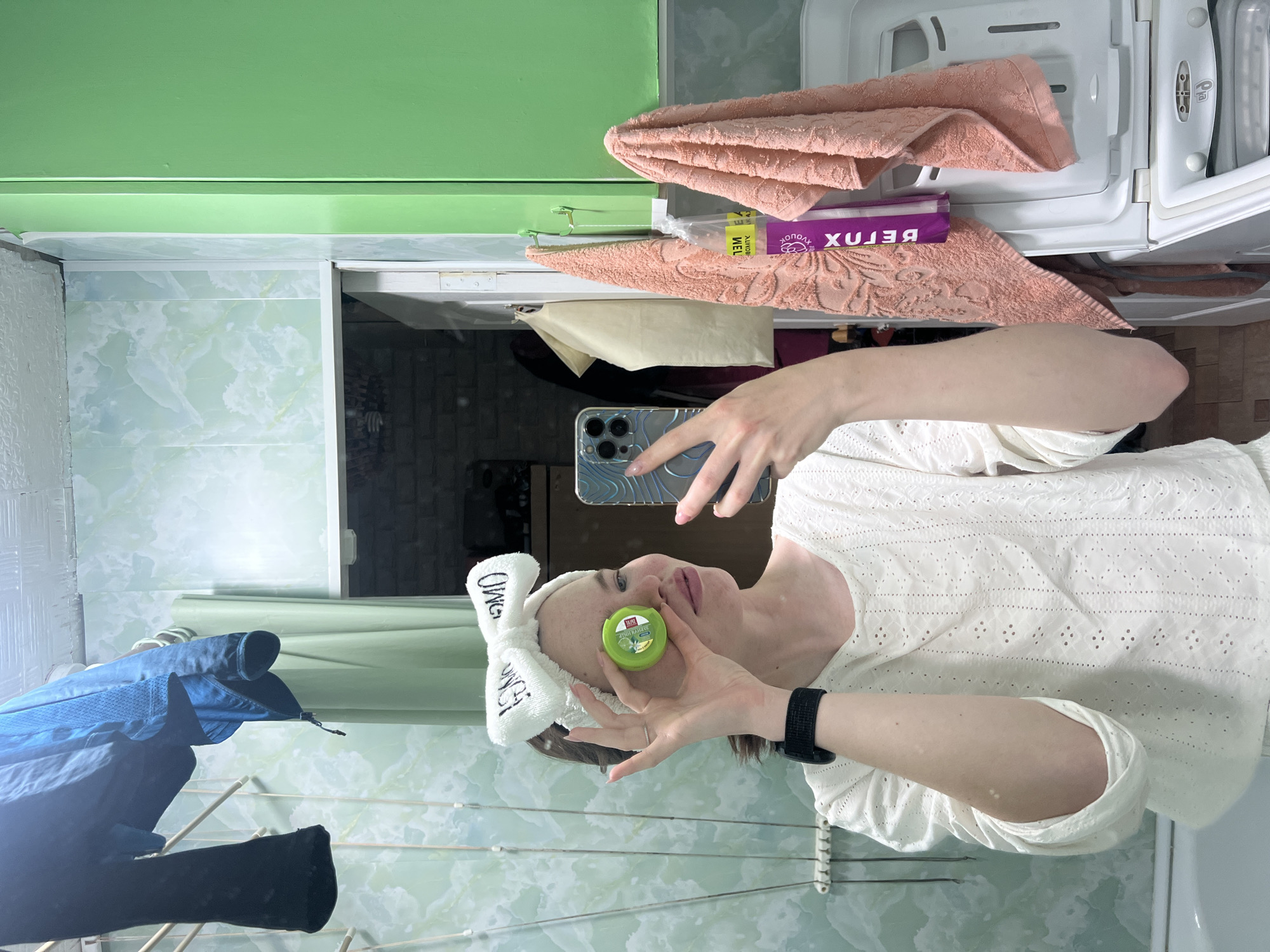 A woman in a white shirt and a white headband with "OMG" written on it is holding a small, round, green container near her face while taking a selfie with her smartphone. She is wearing a black fitness tracker on her wrist. The background is a bathroom with green marble-patterned tiles. A clothes drying rack with blue and black garments hangs on the left side of the image. Above the woman, there's a green cabinet and a washing machine with pink towels and a purple box on top. The location is likely a residential bathroom in Chaikovsky, Russia.