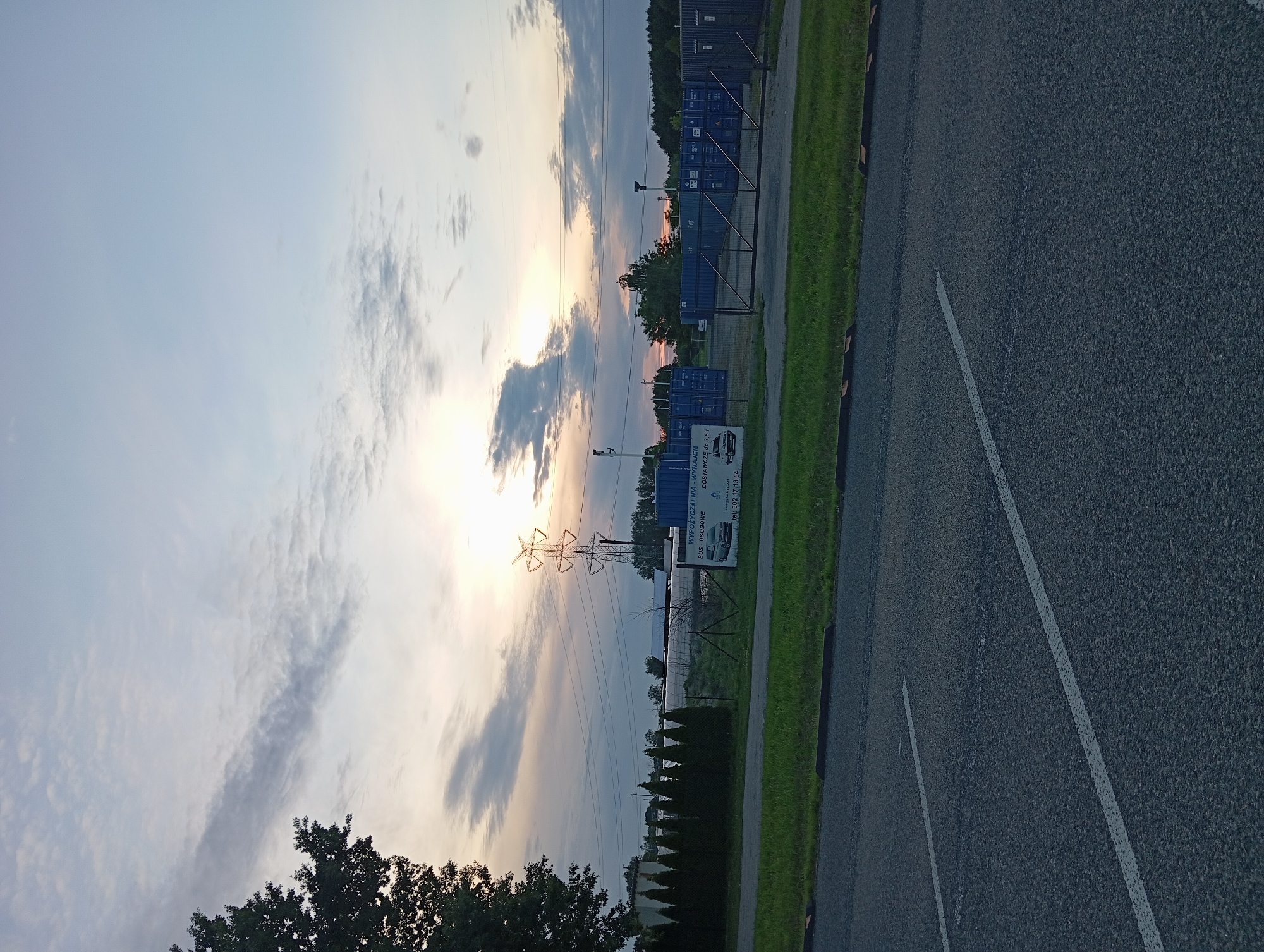 The image depicts an outdoor scene at dusk in Dybów Kolonia, Poland, featuring a road in the foreground and an industrial area with shipping containers in the background. The sky is a mix of blue and grey clouds, with the sun low on the horizon, casting a warm glow. A tall electricity pylon stands prominently in the middle ground.
In the foreground, there is an asphalt road with visible white lane markings. A curb separates the road from a grassy area.
The middle ground shows a fence surrounding an area filled with blue shipping containers. A white sign with Polish text is attached to the fence, advertising "WYPOŻYCZALNIA - WYNAJEM BUS-OSOBOWE DOSTAWCZE DO 3,5T" (Rental - Rental of Buses-Cars Delivery up to 3.5 tons). Below this text, a phone number is visible: "602 17 13 64". Adjacent to the fence, a row of dark green conical trees is planted.
The background consists of a line of trees against the horizon and more of the shipping containers. The overall impression is of an industrial site near a transportation route, with the natural elements of trees and sky creating a serene, yet functional, atmosphere. There are no people or animals visible in the image. The scene appears to be static, with no apparent activity or action.