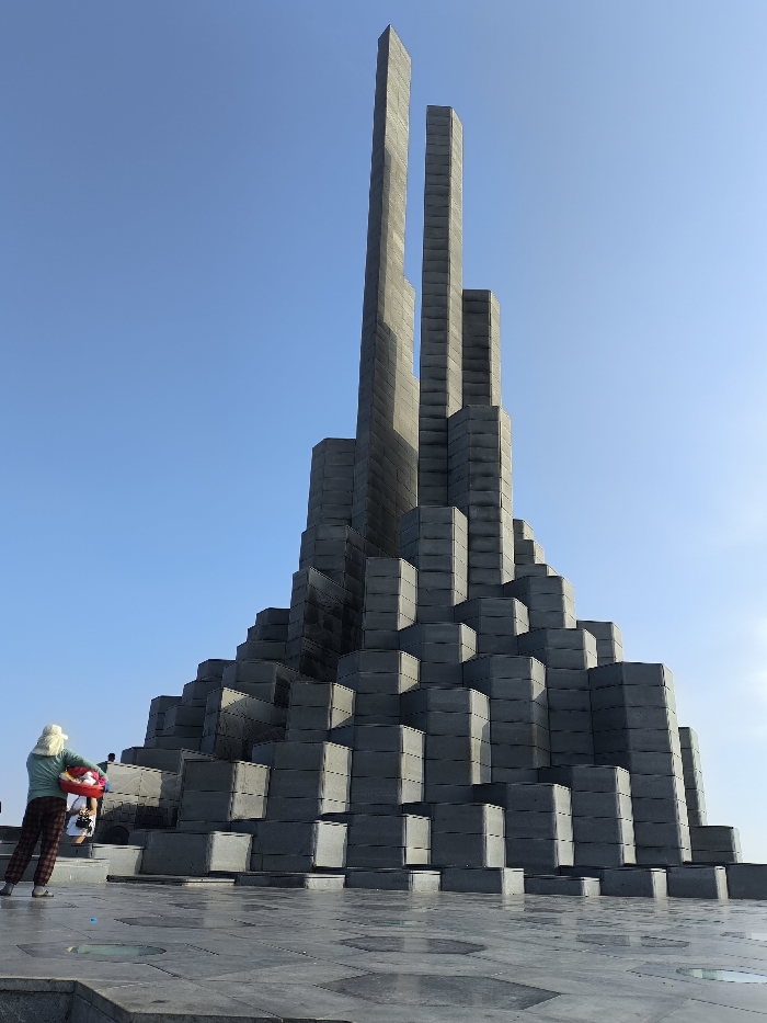 The image displays the Nghinh Phong Tower monument under a clear blue sky in Thành phố Tuy Hòa, Vietnam. The monument, constructed from dark grey, stacked, rectangular block-like components, rises from a wide, tiled plaza. Its base features a stepped, tapering design, culminating in multiple vertical pillars, with two prominent central pillars appearing vertically split near their apex. The ground is paved with dark grey hexagonal and rectangular tiles, featuring embedded circular clear panels that reflect light.
One individual is visible in the lower-left foreground, facing away from the camera, observing the monument. This person wears a light-colored hat, a light green long-sleeved shirt, and dark plaid trousers. They are holding a red and white inflatable ring. A second person, partially obscured behind the first, is visible in a white shirt. The scene is brightly lit by natural sunlight, casting subtle shadows.