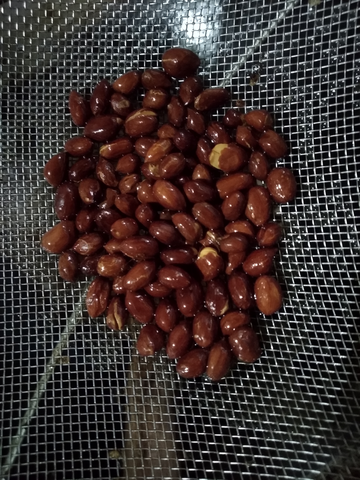 A close-up, overhead shot shows a pile of roasted peanuts resting in a metal colander. The peanuts are dark brown, with some showing lighter, creamy-colored centers. They are clustered together, filling most of the colander's surface. The colander itself is made of a fine, silver mesh with a grid pattern, and it is placed on a dark, unidentifiable surface. No people or environmental context beyond the colander and its contents are visible. There is no indication of the location being Balaraja, Indonesia.