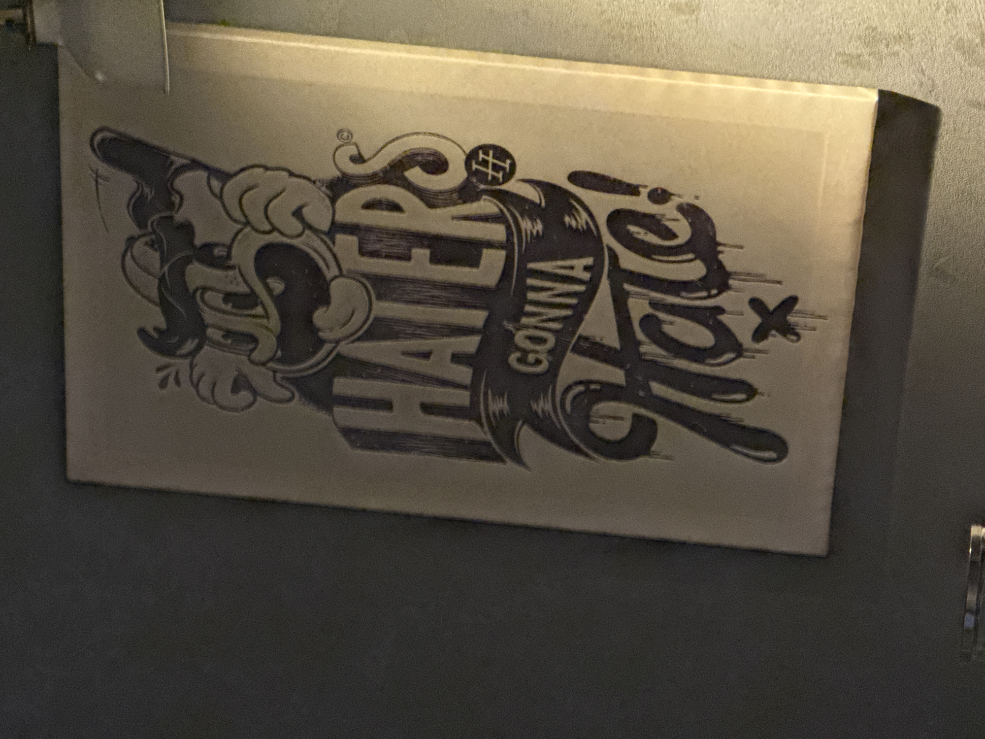 A close-up, angled shot shows a motivational poster or artwork featuring a cartoon character with a large head and a wide smile. The character appears to be holding a baseball bat. The text on the poster reads "HATERS GONNA HATE!" in a stylized font with dripping effects. There is a small emblem with "HH" inside a circle to the right of the word "HATERS." The artwork is predominantly in dark purple and white tones on a beige background. The poster is hanging on a dark gray wall, casting a shadow on the right side. The lighting suggests an indoor setting, possibly a restaurant or bar, with a light fixture visible in the upper right corner. The image does not provide any information about the city of Chaikovsky, Russia.